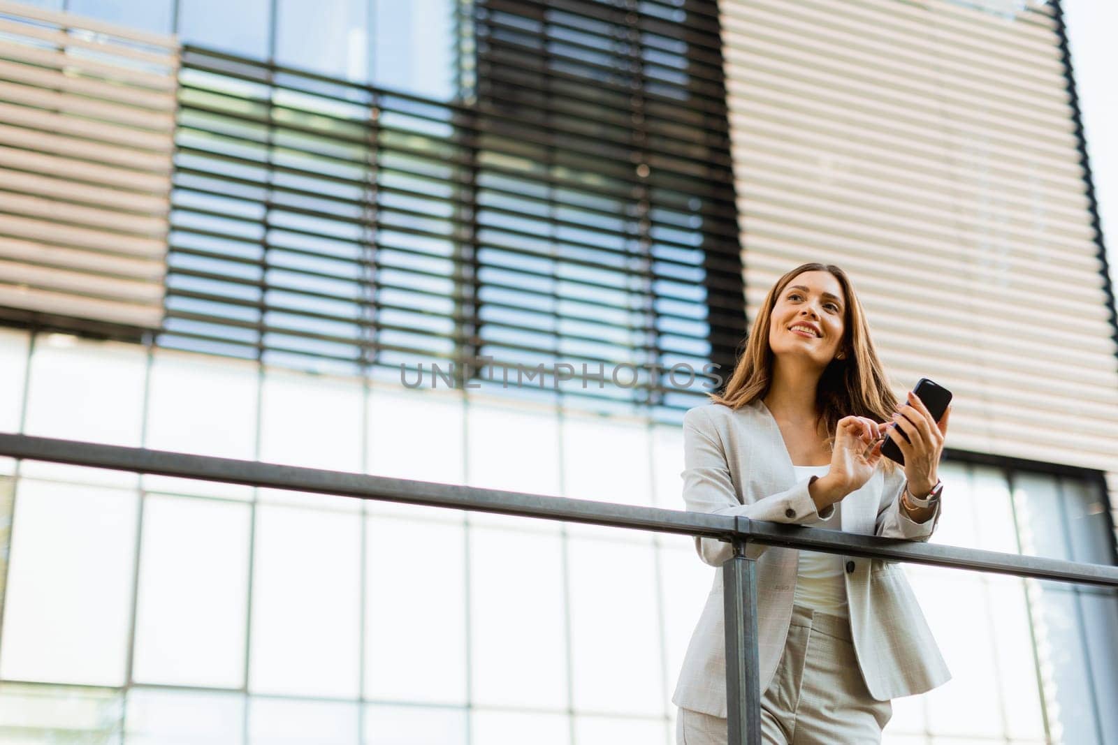 Cheerful business woman stands on a balcony, engaged with her smartphone against a backdrop of modern architecture, soaking in the warm sunlight and vibrant atmosphere