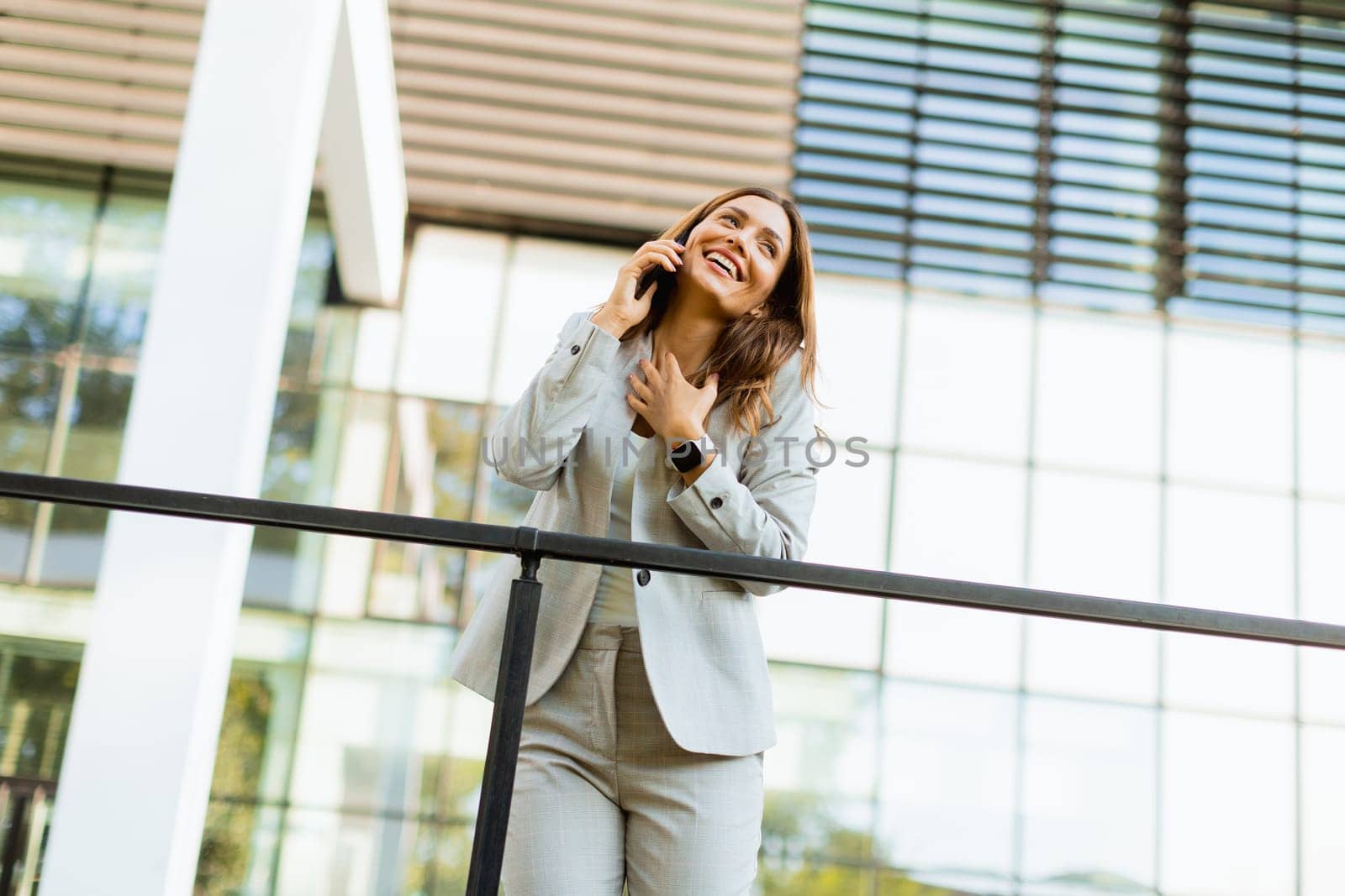 Joyful business woman celebrates a moment outdoors, engaging in a cheerful phone conversation while leaning against a railing beside an elegant building.