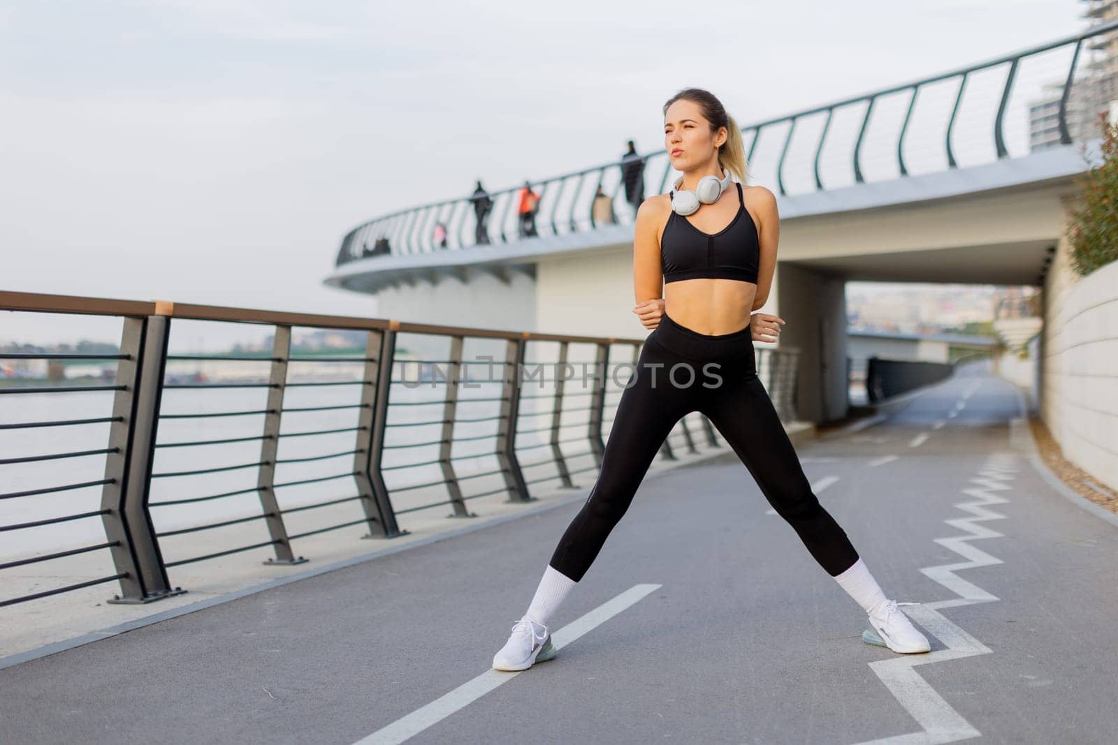A fit woman wearing athletic clothing performs a stretching routine on a riverside pathway, with serene water views and leisurely walkers enjoying a peaceful morning outside.