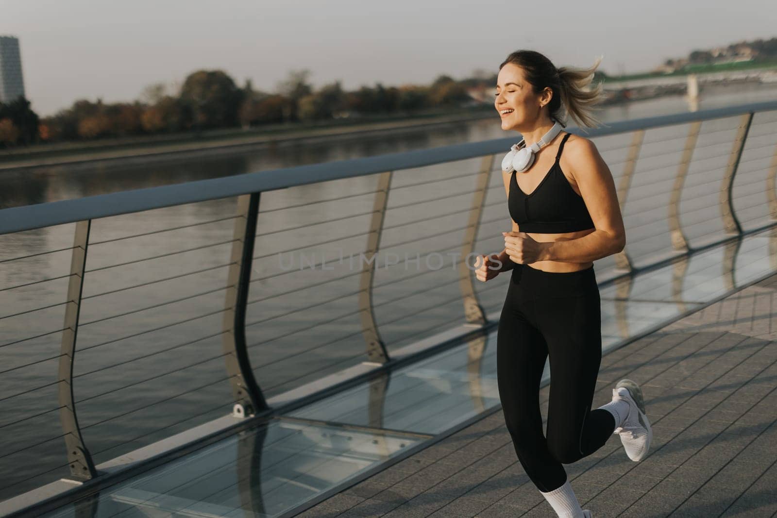 A young woman in athletic attire runs energetically along a riverside path, embracing the serene atmosphere of a beautiful morning, fueled by her favorite music.