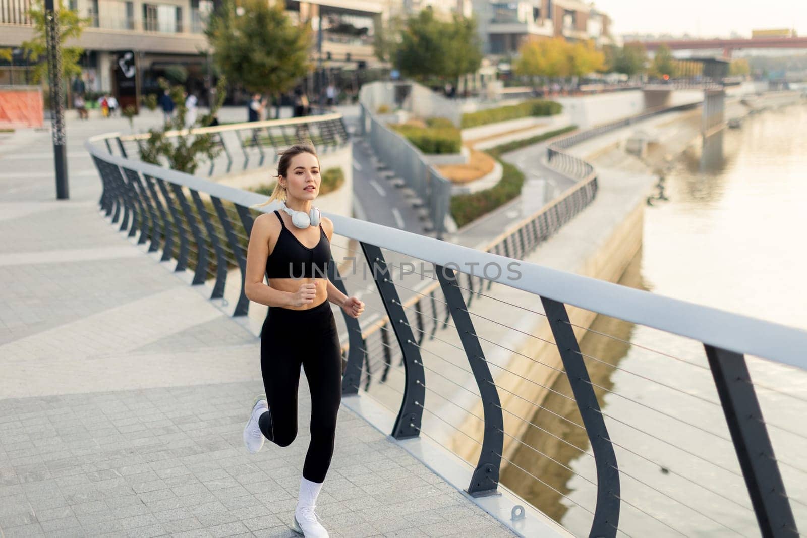 A dedicated runner maintains her pace on a riverwalk surrounded by urban architecture and lush greenery, embracing a beautiful morning and an active lifestyle.
