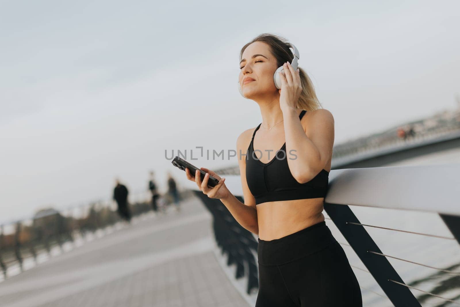 A young woman stands relaxed on a bridge, headphones on, savoring music while basking in the gentle glow of sunset near the water's edge.