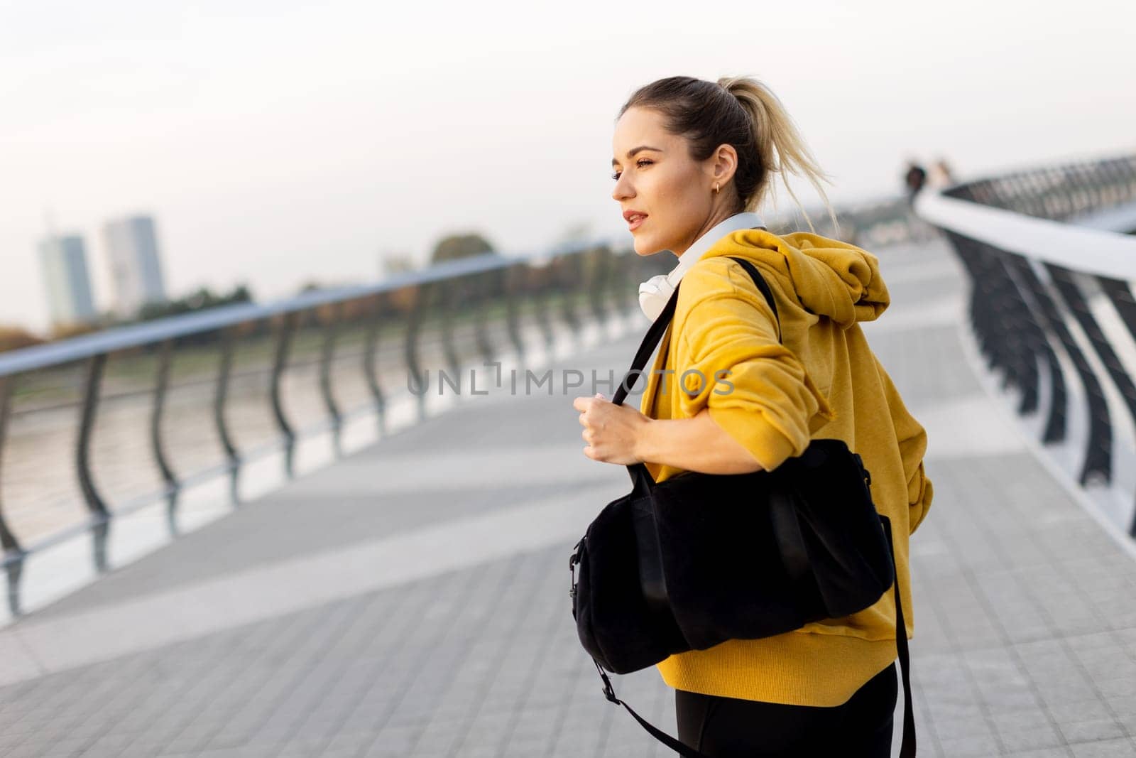 Walking along the riverside path, a cheerful woman in a yellow hoodie beams with joy. The evening sky casts a warm glow, creating a serene atmosphere for a leisurely stroll