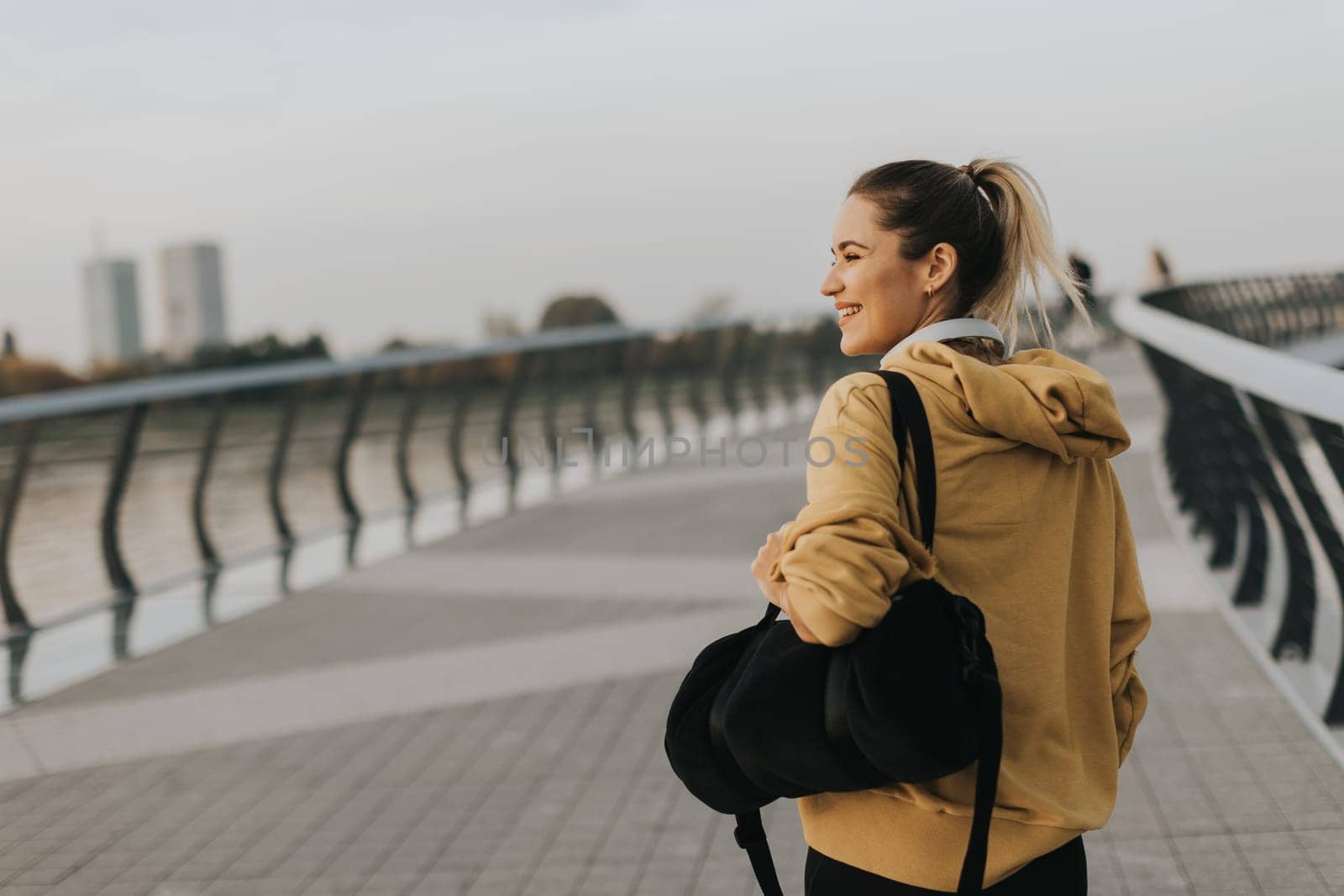 With a joyful smile, a young woman in a yellow hoodie strolls along a scenic riverside path, her black bag slung over her shoulder, embracing the gentle evening breeze.