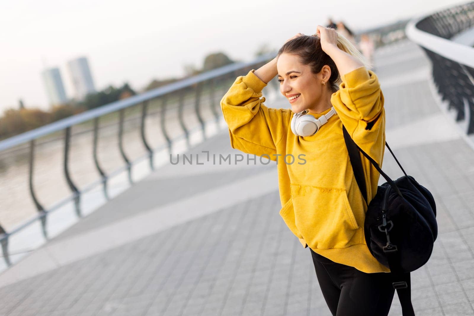 A cheerful woman strolls along a picturesque waterfront pathway, wearing a bright yellow hoodie and headphones. Her vibrant smile reflects the sunny ambiance of the day.