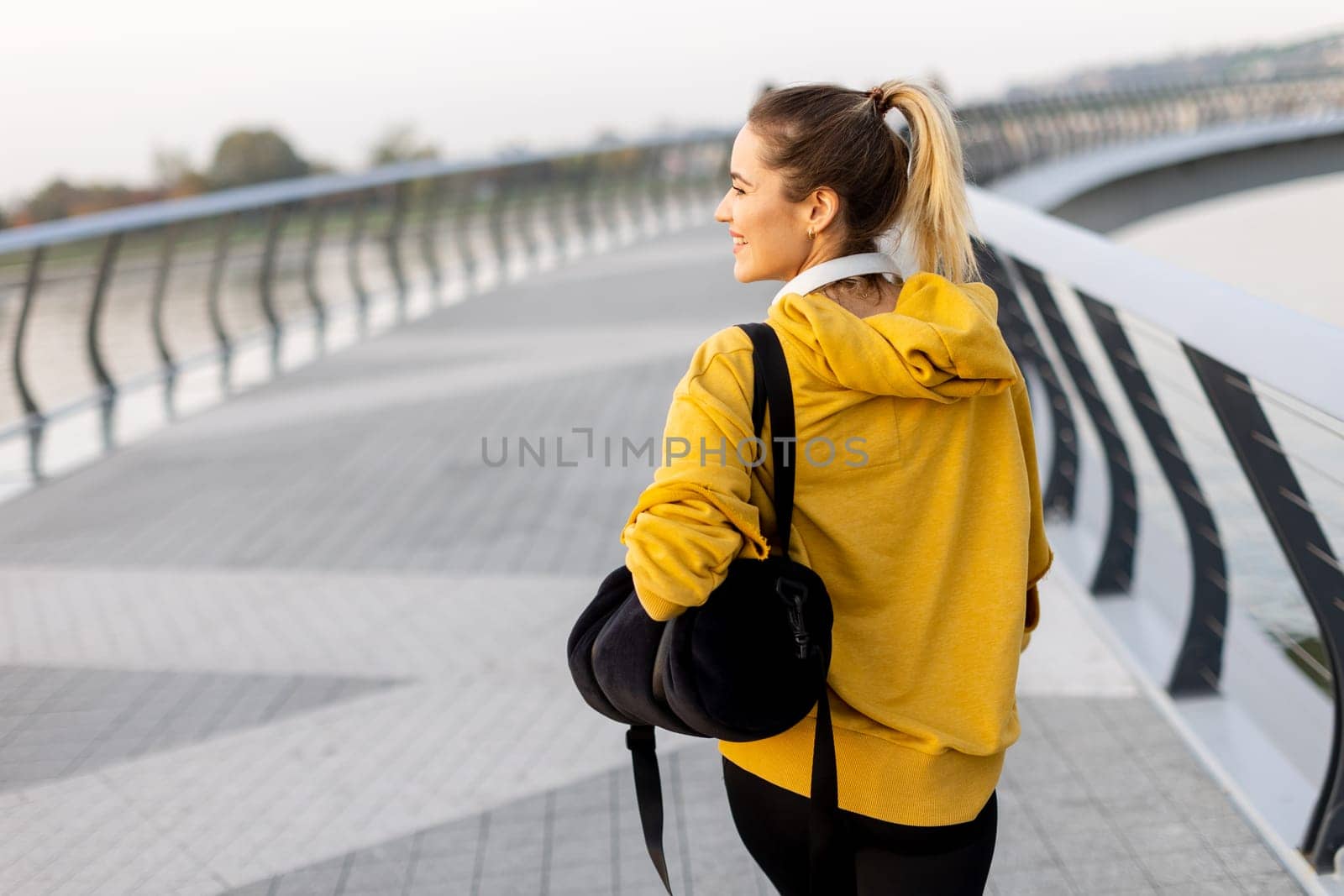 A carefree woman walks along a contemporary riverside path, her cheerful demeanor captured as she enjoys the picturesque sunset and fresh air.