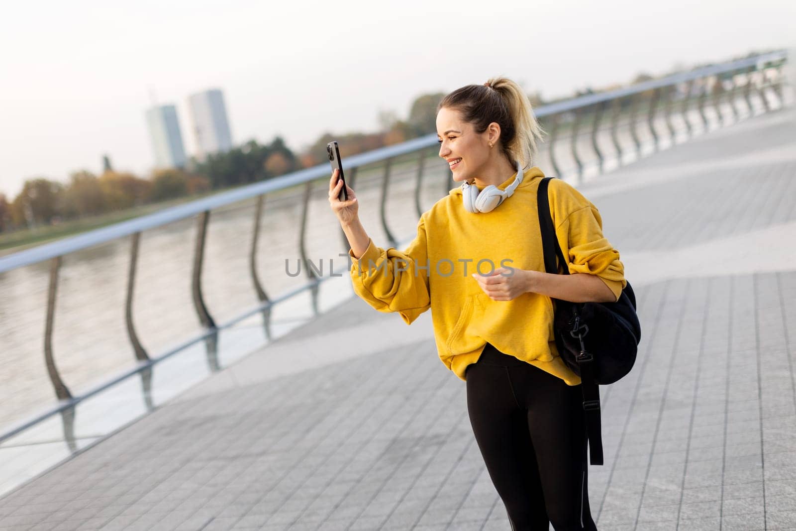 Joyful young woman strolls along a riverside walkway, engaged in a lively phone conversation while wearing a bright yellow sweatshirt and carrying a black bag