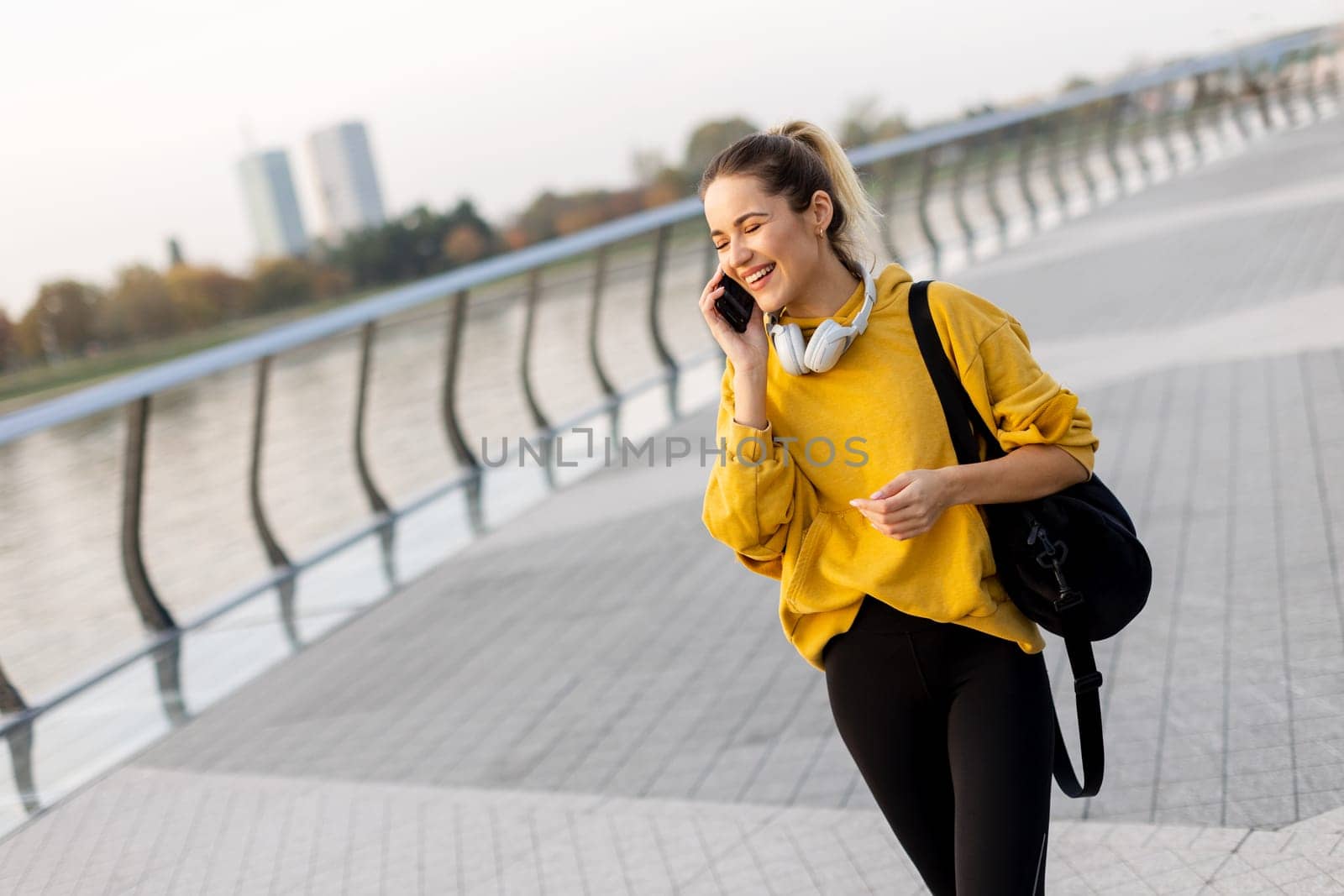 A cheerful woman dressed in a vibrant yellow sweater laughs on her phone while strolling along a picturesque riverside walkway, enjoying the warm sunshine.