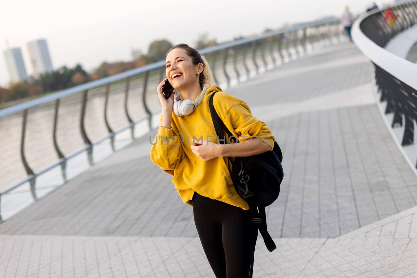 A cheerful woman dressed in a vibrant yellow sweater laughs on her phone while strolling along a picturesque riverside walkway, enjoying the warm sunshine.