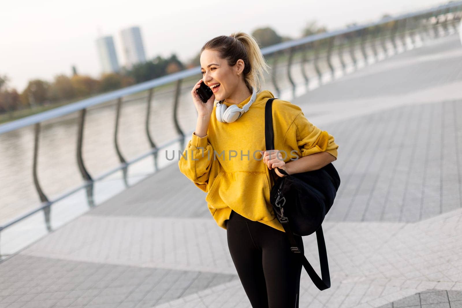 Joyful young woman strolls along a riverside walkway, engaged in a lively phone conversation while wearing a bright yellow sweatshirt and carrying a black bag