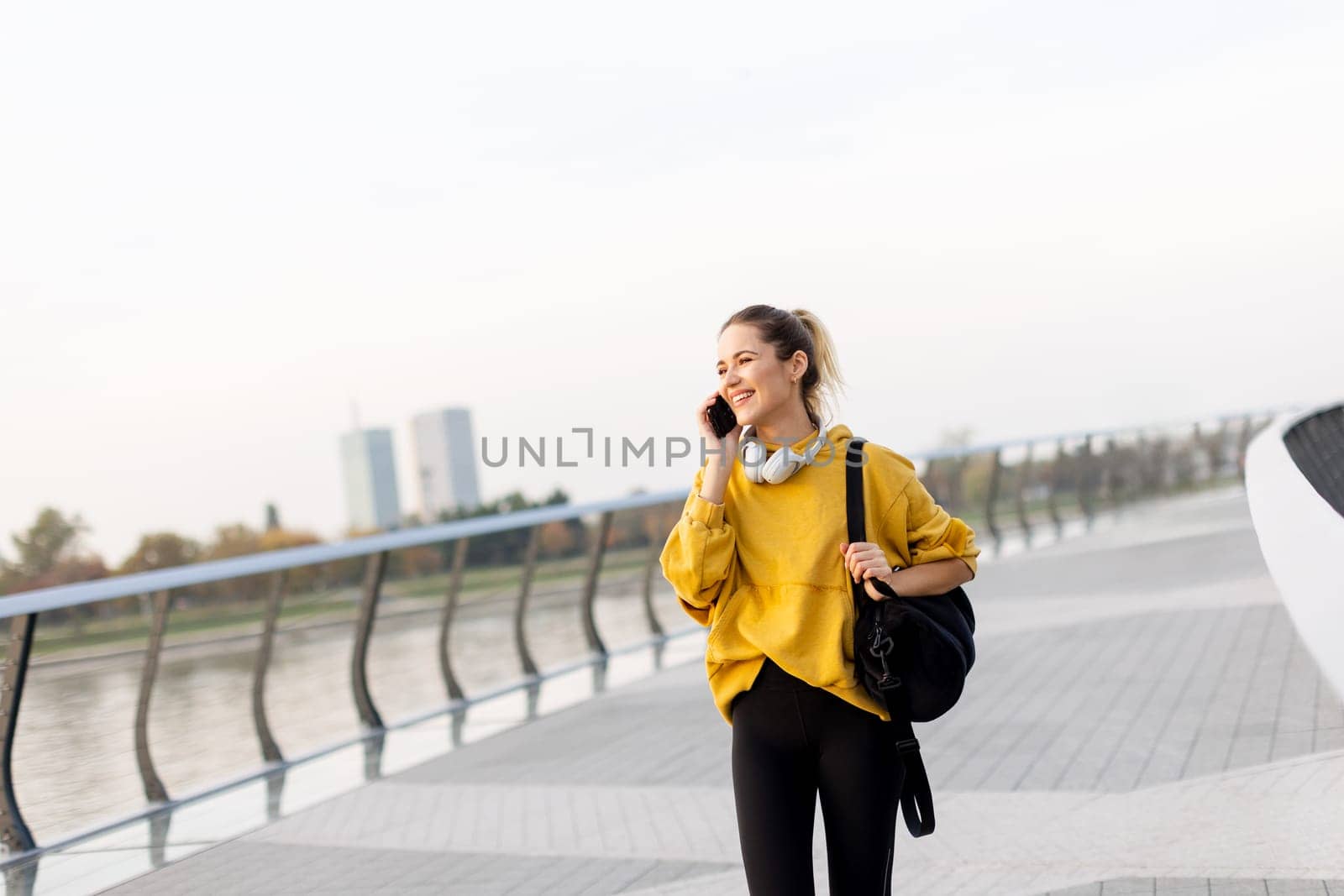 Joyful young woman strolls along a riverside walkway, engaged in a lively phone conversation while wearing a bright yellow sweatshirt and carrying a black bag
