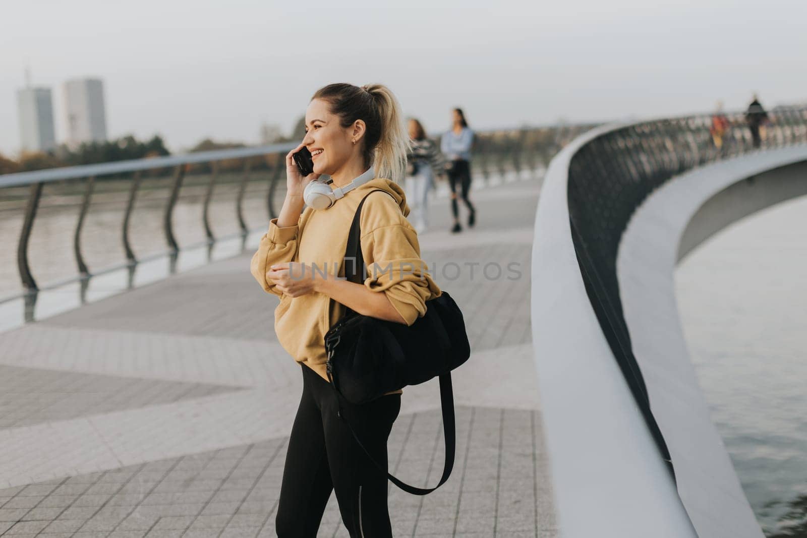 As the sun rises over the skyline, a young woman dressed in a vibrant yellow sweater walks alongside the river, captivated by her smartphone while listening to music.