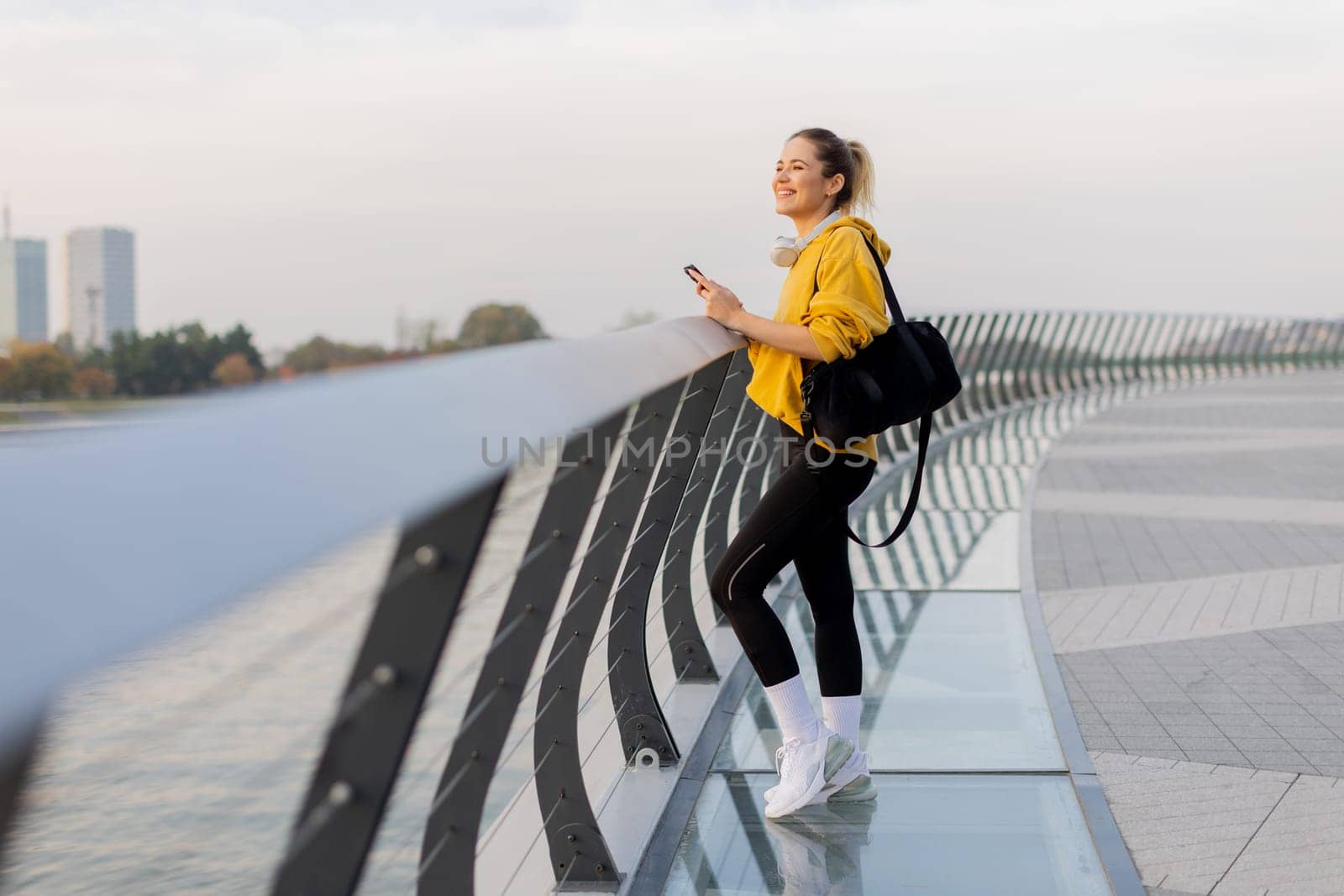 A young woman wearing a bright yellow jacket leans against a modern railing as she focuses on her phone, enjoying a tranquil riverside stroll on a pleasant afternoon.