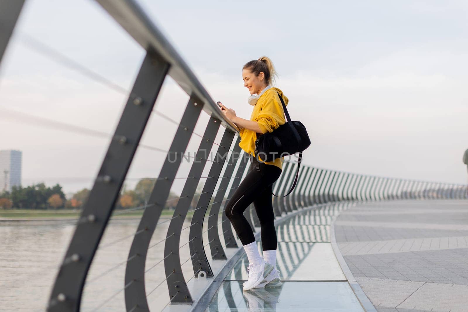 A young woman wearing a bright yellow jacket leans against a modern railing as she focuses on her phone, enjoying a tranquil riverside stroll on a pleasant afternoon.