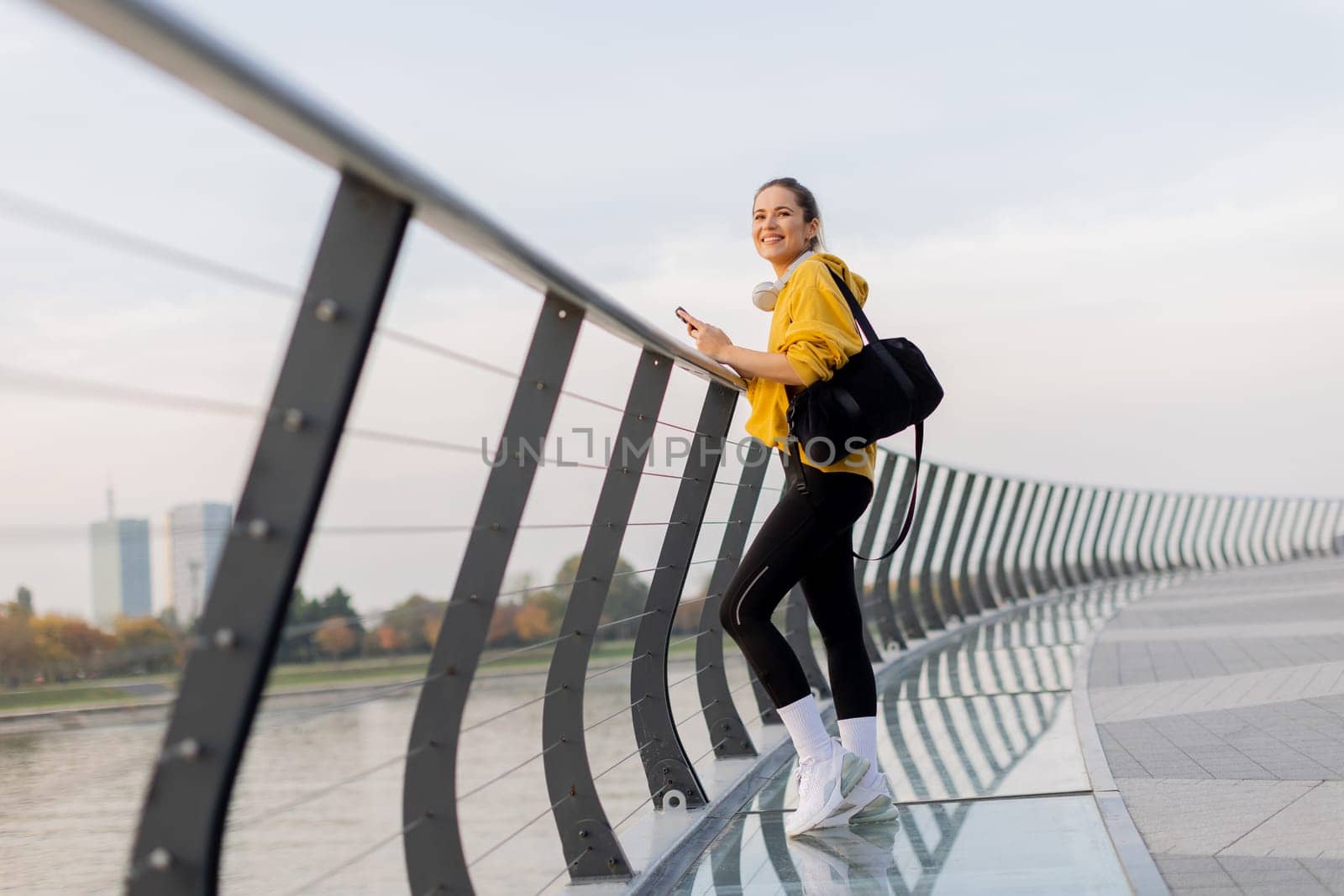 Young woman with a confident smile strolls along a sleek, glass railing by the river, dressed comfortably for an afternoon of leisure in a bright yellow hoodie and leggings.