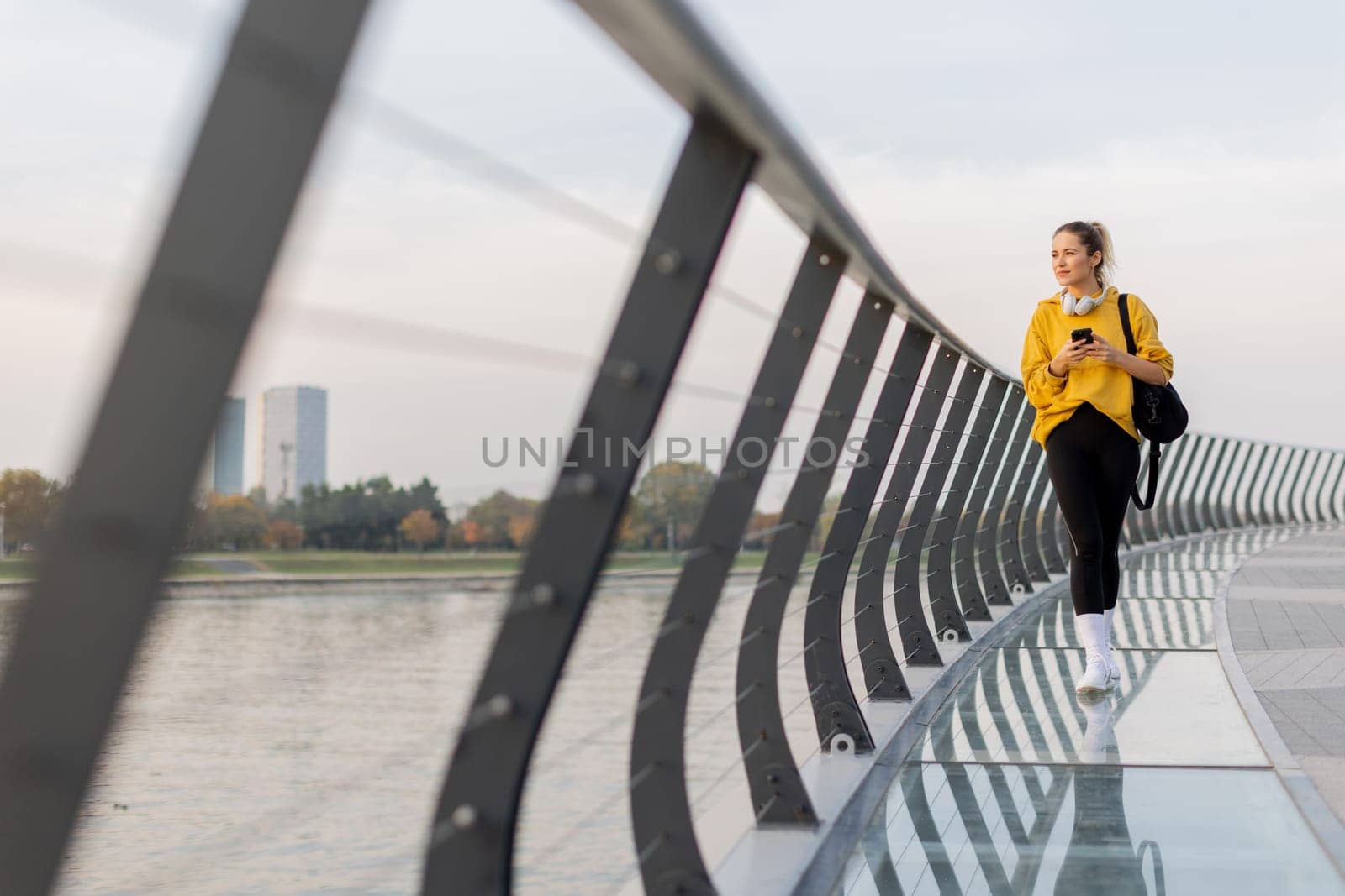 A young woman wearing a bright yellow jacket leans against a modern railing as she focuses on her phone, enjoying a tranquil riverside stroll on a pleasant afternoon.