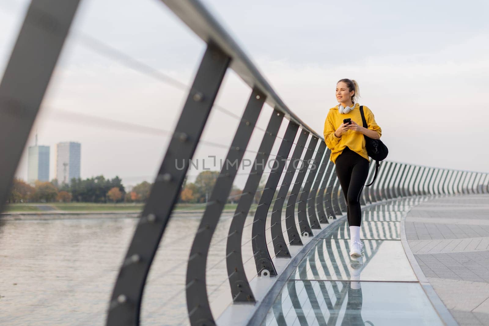 A young woman wearing a bright yellow jacket leans against a modern railing as she focuses on her phone, enjoying a tranquil riverside stroll on a pleasant afternoon.