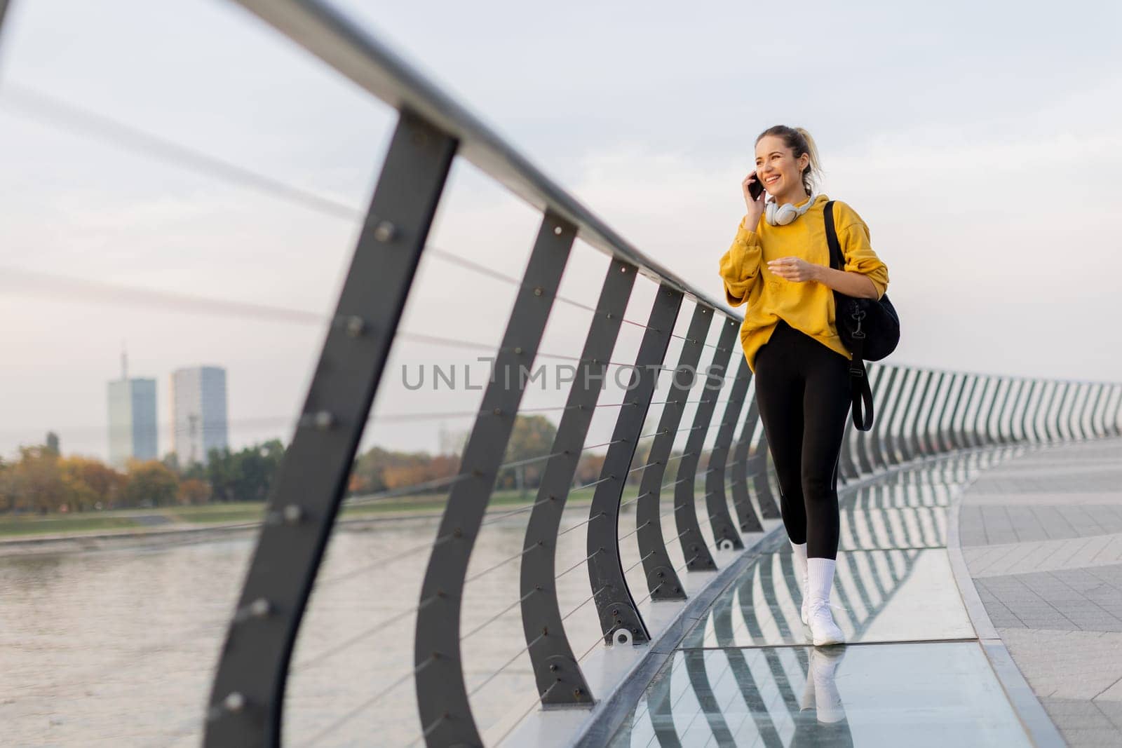Young woman walks casually along a sleek pedestrian bridge, talking on her phone. The tranquil waters reflect a picturesque urban skyline as twilight approaches
