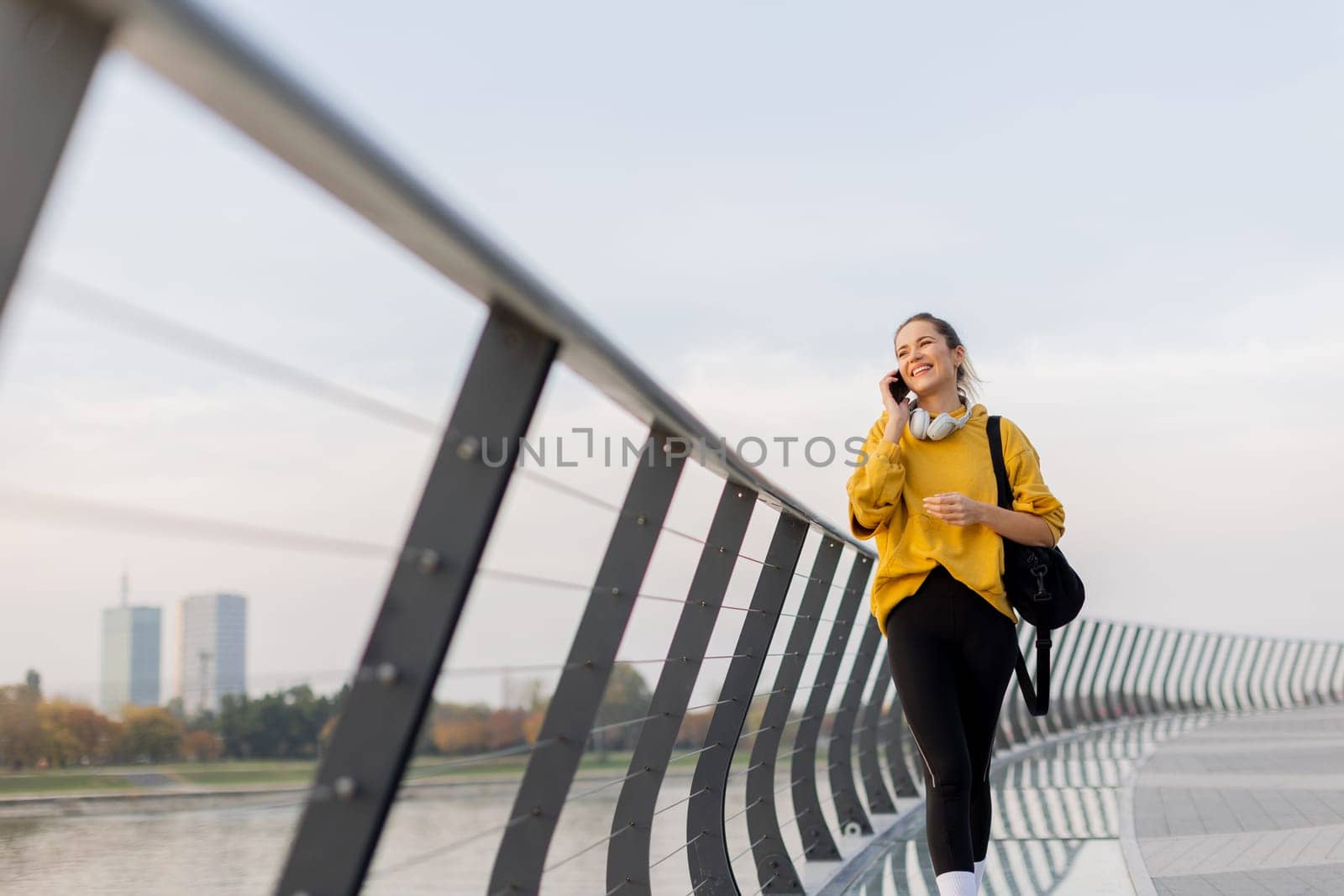Young woman walks casually along a sleek pedestrian bridge, talking on her phone. The tranquil waters reflect a picturesque urban skyline as twilight approaches