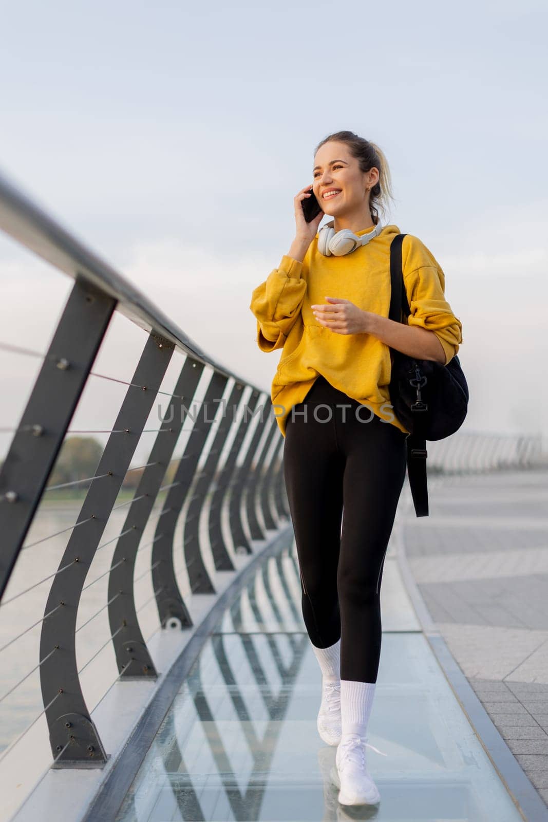 Young woman walks casually along a sleek pedestrian bridge, talking on her phone. The tranquil waters reflect a picturesque urban skyline as twilight approaches