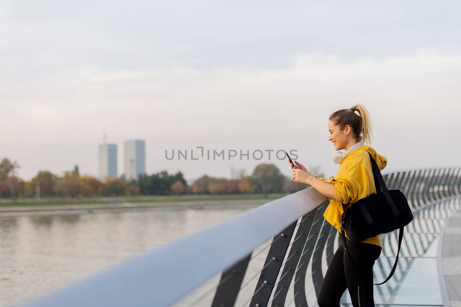 Young woman with a confident smile strolls along a sleek, glass railing by the river, dressed comfortably for an afternoon of leisure in a bright yellow hoodie and leggings.