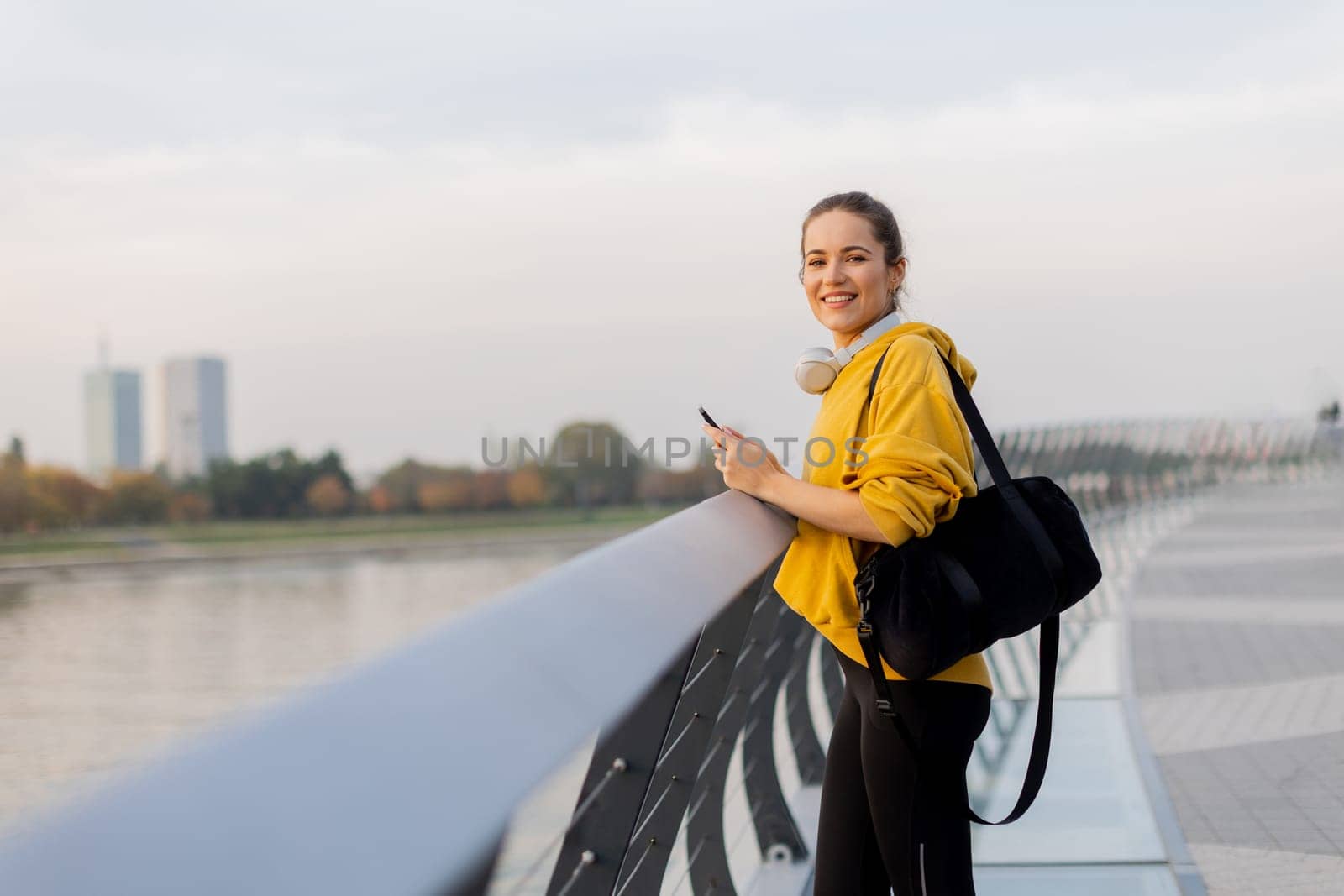 Young woman with a confident smile strolls along a sleek, glass railing by the river, dressed comfortably for an afternoon of leisure in a bright yellow hoodie and leggings.