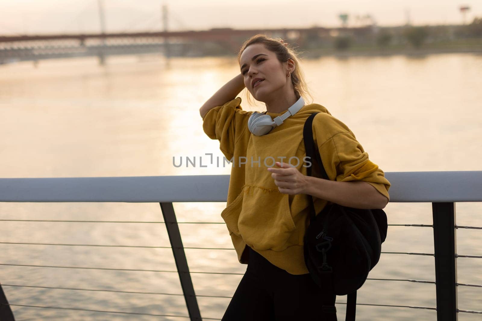 With the sun setting behind her, a cheerful young woman stands by the water, savoring a tranquil moment as she playfully pulls back her hair, feeling carefree and content