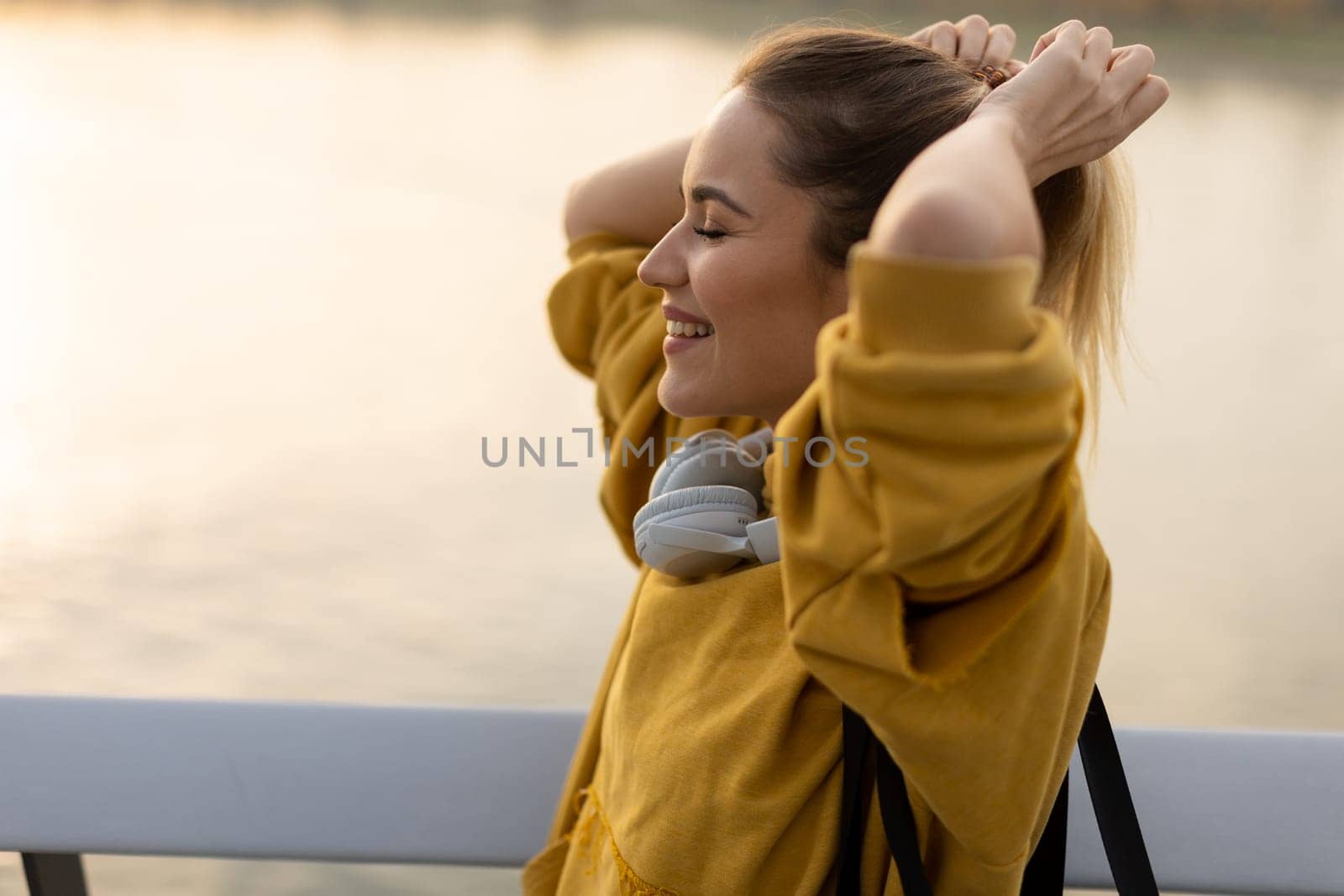 With the sun setting behind her, a cheerful young woman stands by the water, savoring a tranquil moment as she playfully pulls back her hair, feeling carefree and content