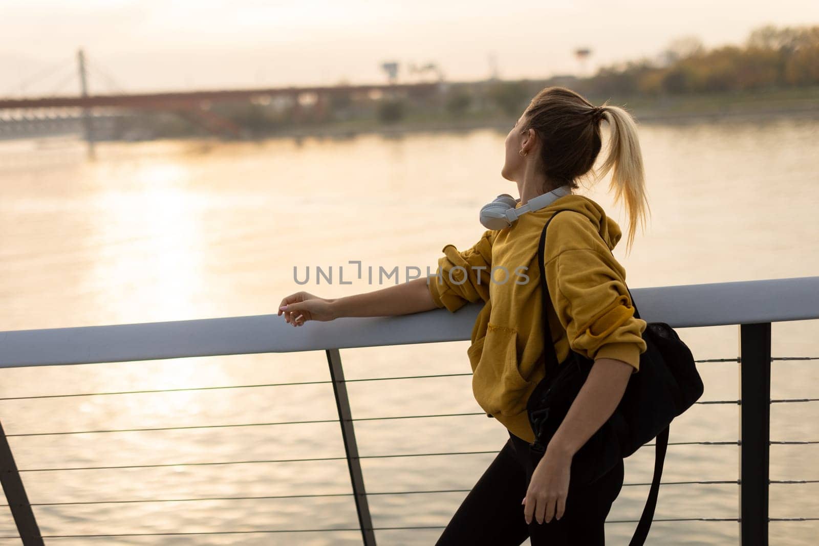 A carefree woman walks along a contemporary riverside path, her cheerful demeanor captured as she enjoys the picturesque sunset and fresh air.
