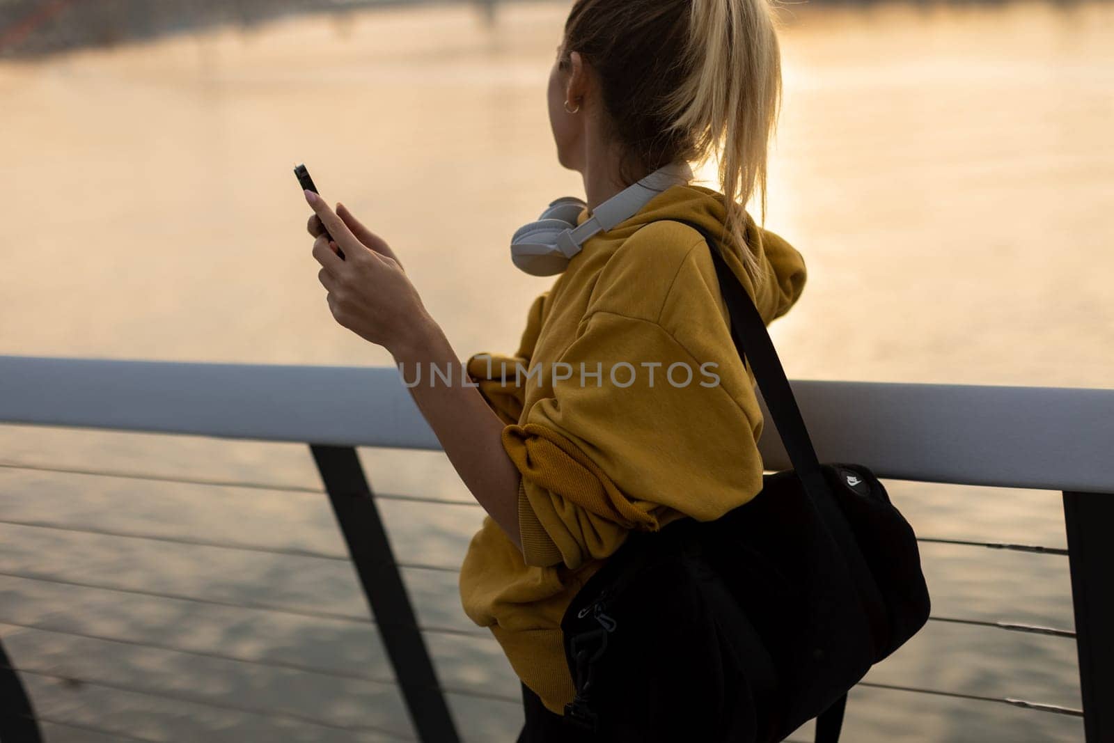 As the sun sets over the tranquil river, a young woman wearing a yellow hoodie and headphones smiles while texting, capturing a moment of joy and relaxation in nature