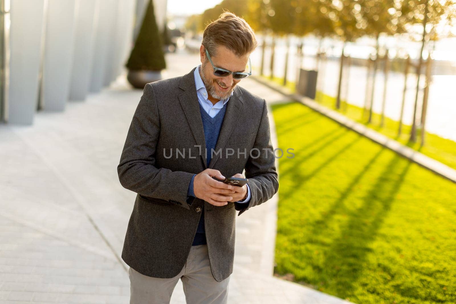 A professional man in stylish attire leisurely walks outside a sleek building, smiling as he interacts with his phone under the warm afternoon sun, exuding confidence.