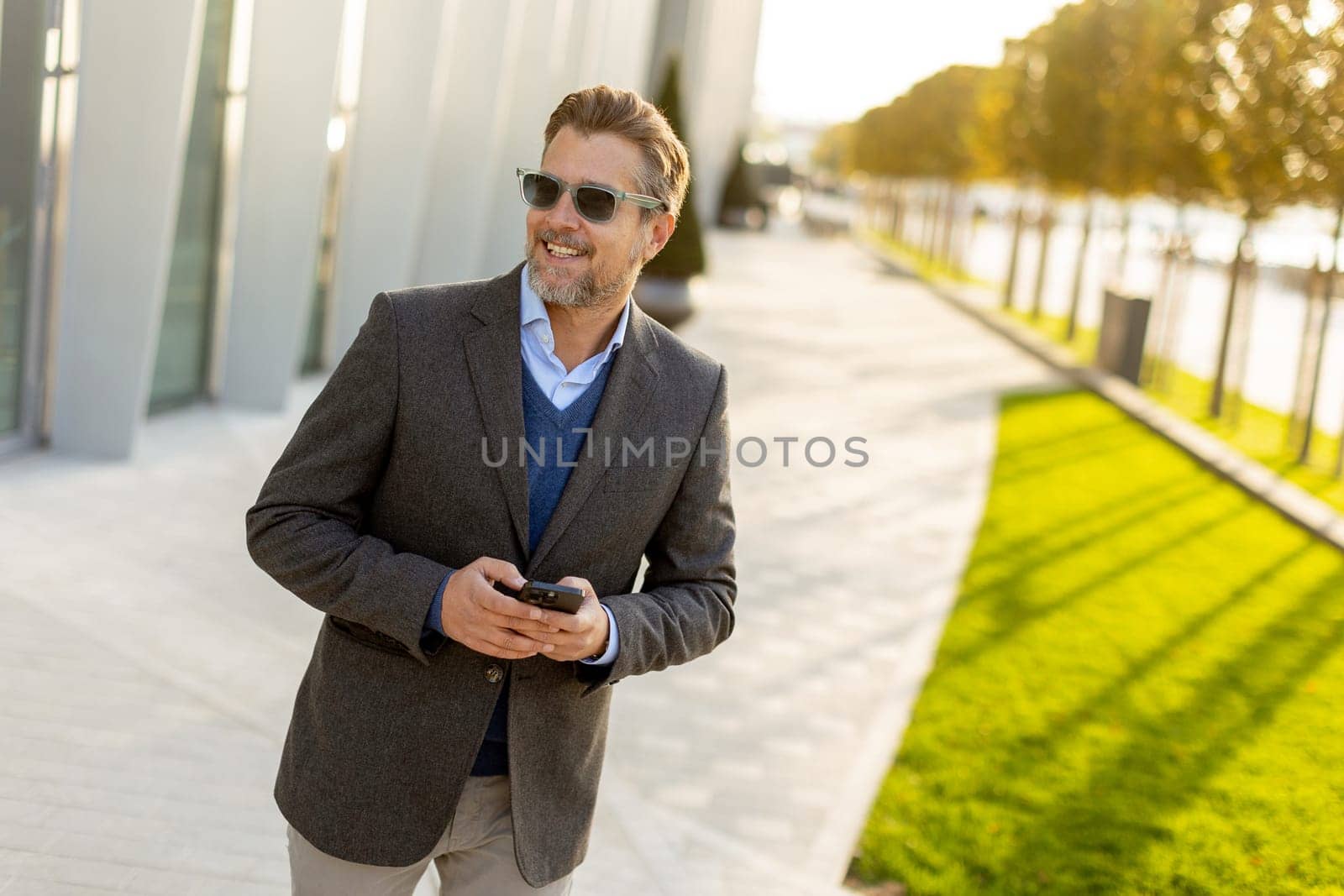 A professional man in stylish attire leisurely walks outside a sleek building, smiling as he interacts with his phone under the warm afternoon sun, exuding confidence.