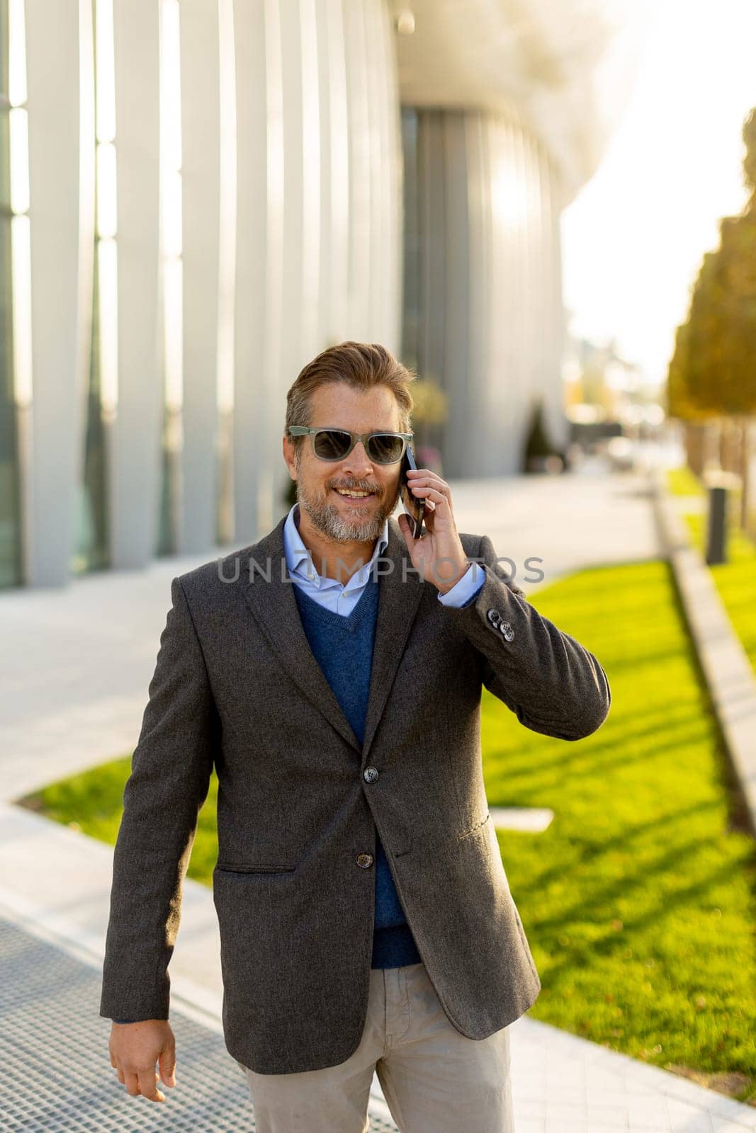 A well-dressed man strolls along a sleek pathway, chatting on his phone under the warm glow of the afternoon sun, surrounded by contemporary architecture and greenery.
