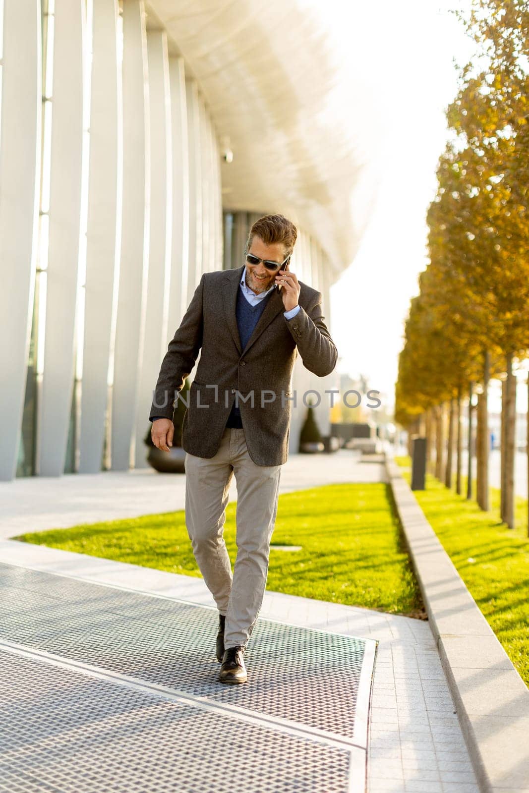 A well-dressed man strolls along a sleek pathway, chatting on his phone under the warm glow of the afternoon sun, surrounded by contemporary architecture and greenery.