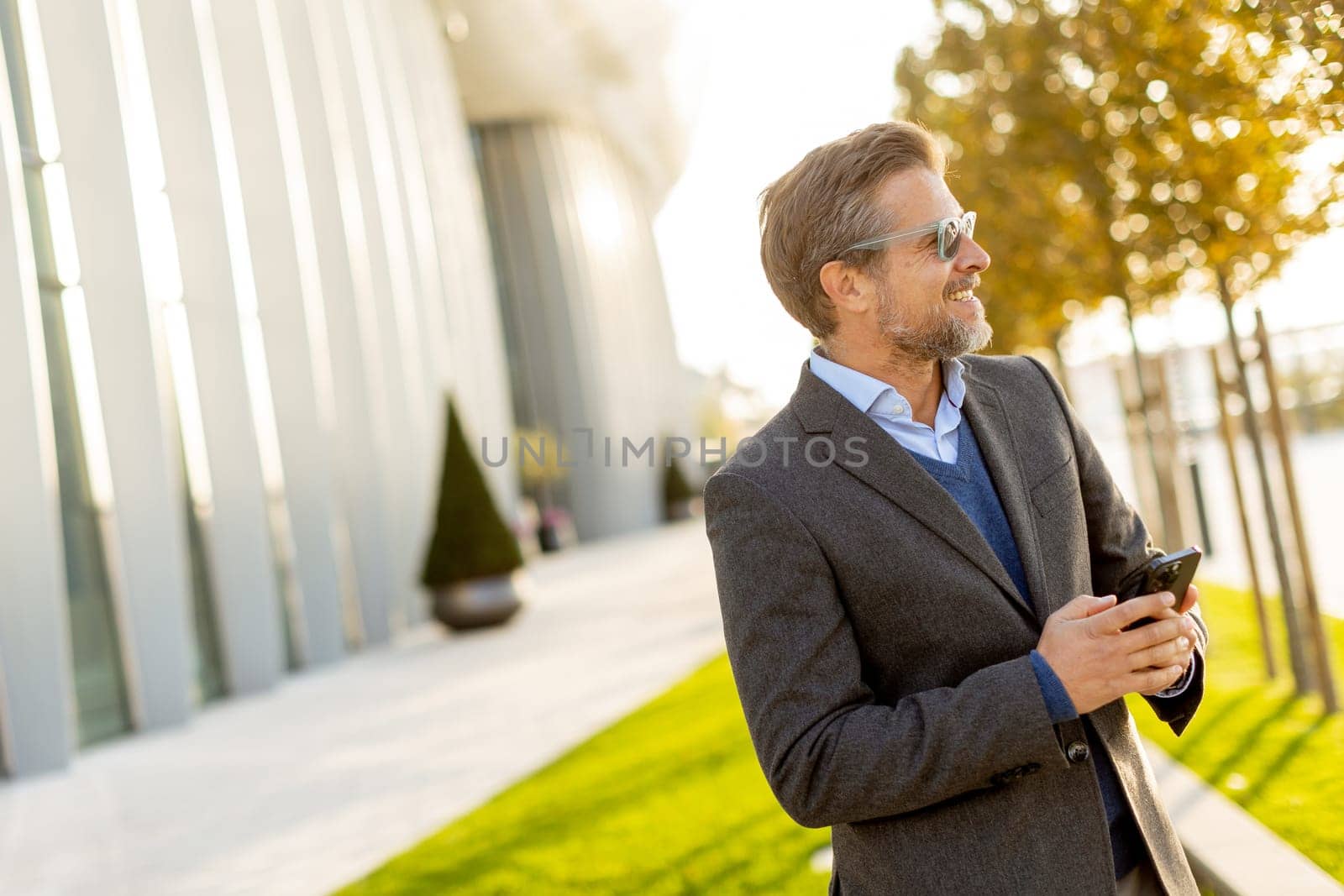 A professional man in stylish attire leisurely walks outside a sleek building, smiling as he interacts with his phone under the warm afternoon sun, exuding confidence.