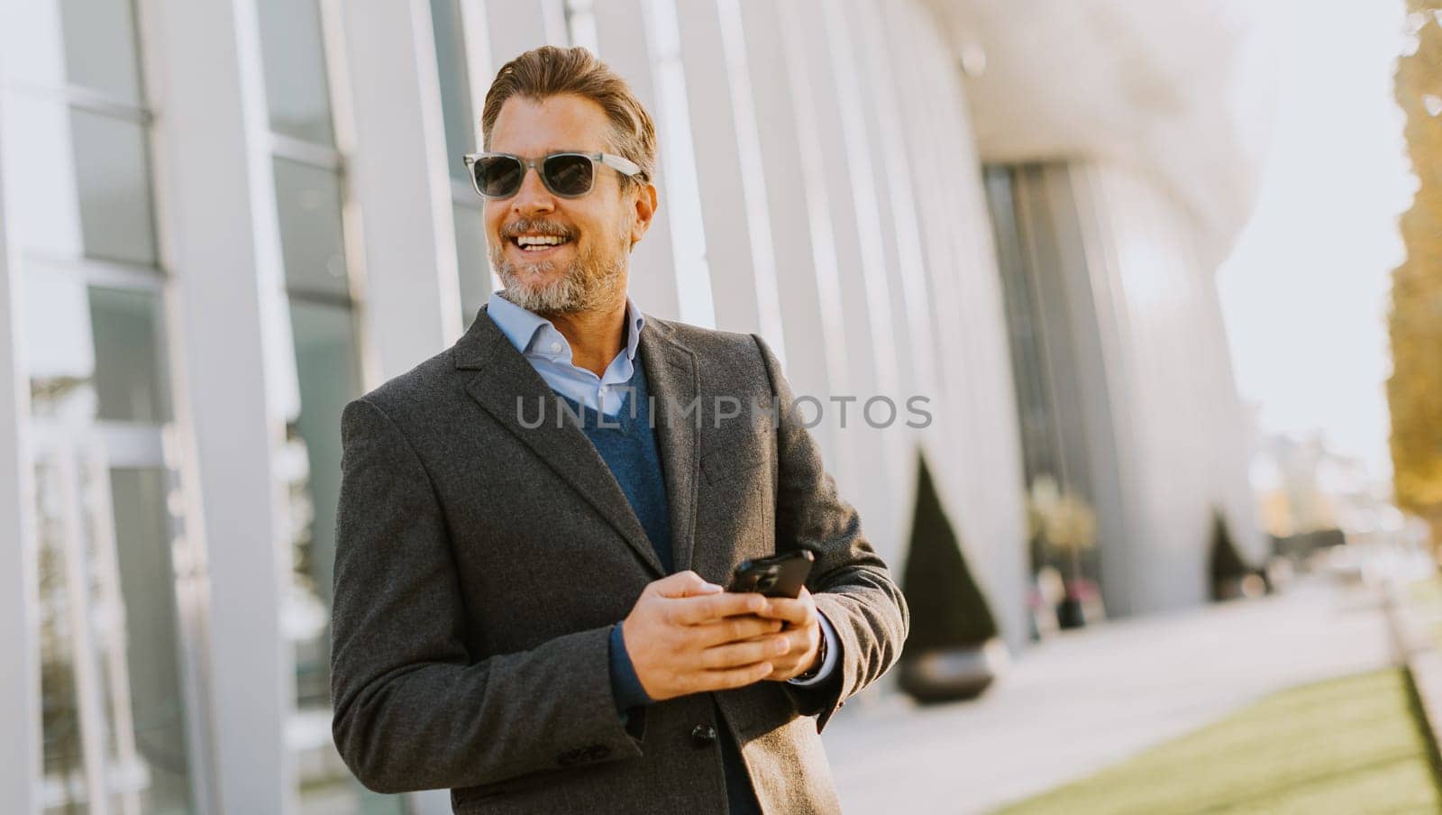 A professional man in stylish attire leisurely walks outside a sleek building, smiling as he interacts with his phone under the warm afternoon sun, exuding confidence.