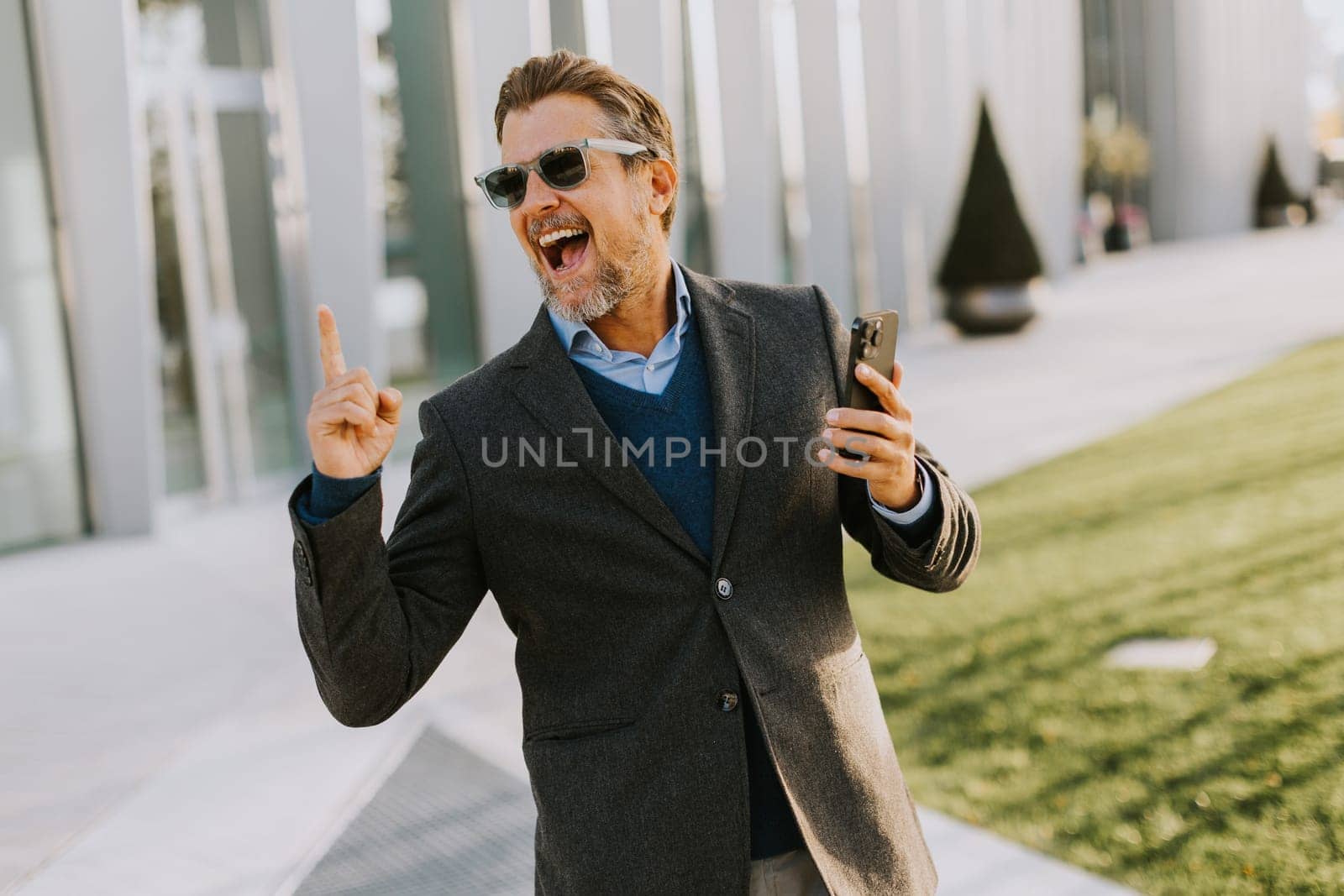 A cheerful man in a fashionable suit and sunglasses laughs heartily, gesturing with excitement while enjoying a lively moment outside in an urban landscape.