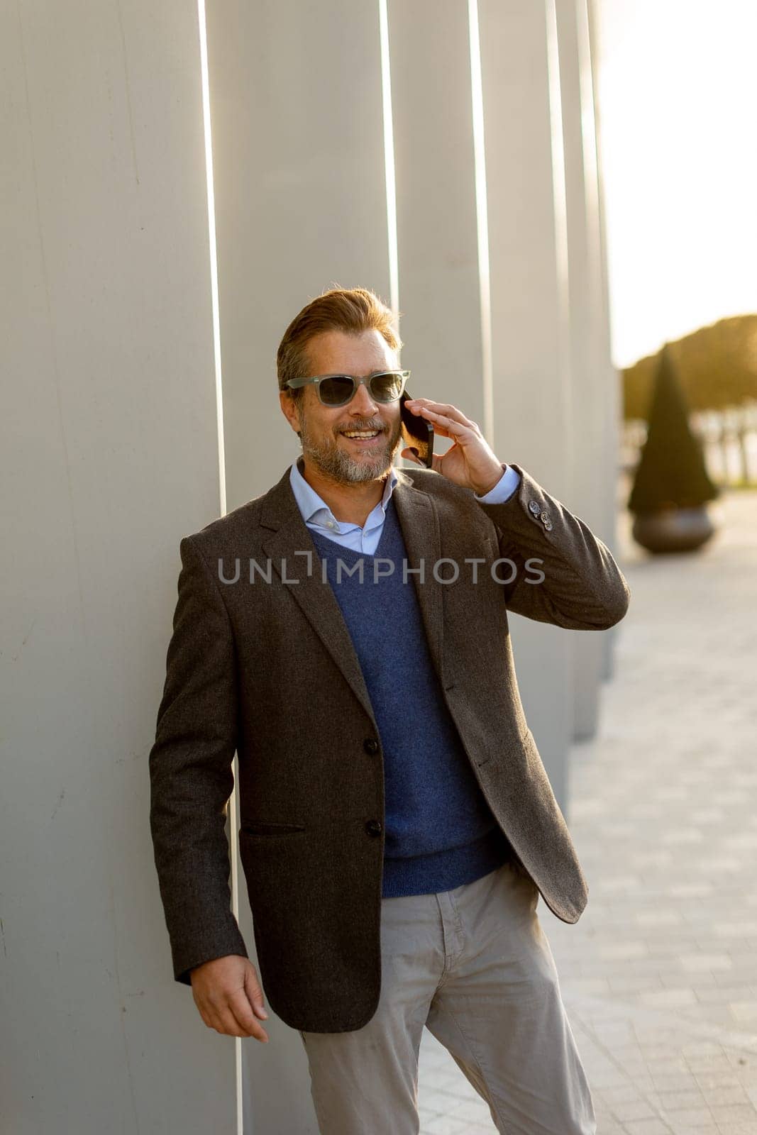 A well-dressed man is leaning casually against a sleek white wall while talking on his phone. The warm light of sunset beautifully highlights the modern architecture around him.