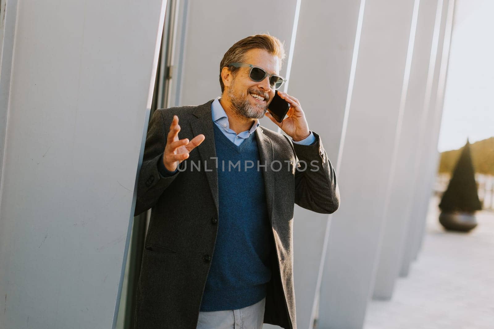 A well-dressed man enjoys a casual conversation on the phone while walking along a sunlit pathway next to contemporary buildings, creating an ambiance of sophistication.