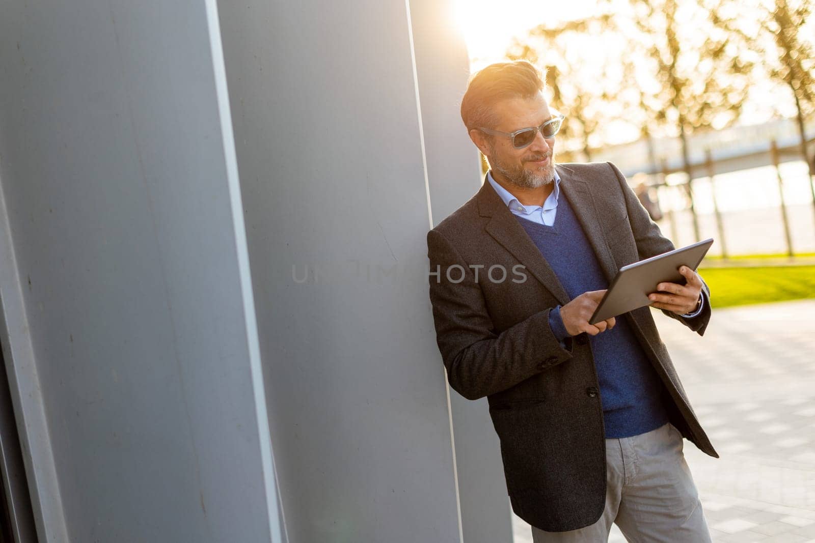 A man in a tailored suit stands confidently against a sleek wall, engrossed in his tablet as the sun illuminates the park around him, creating a joyful atmosphere.