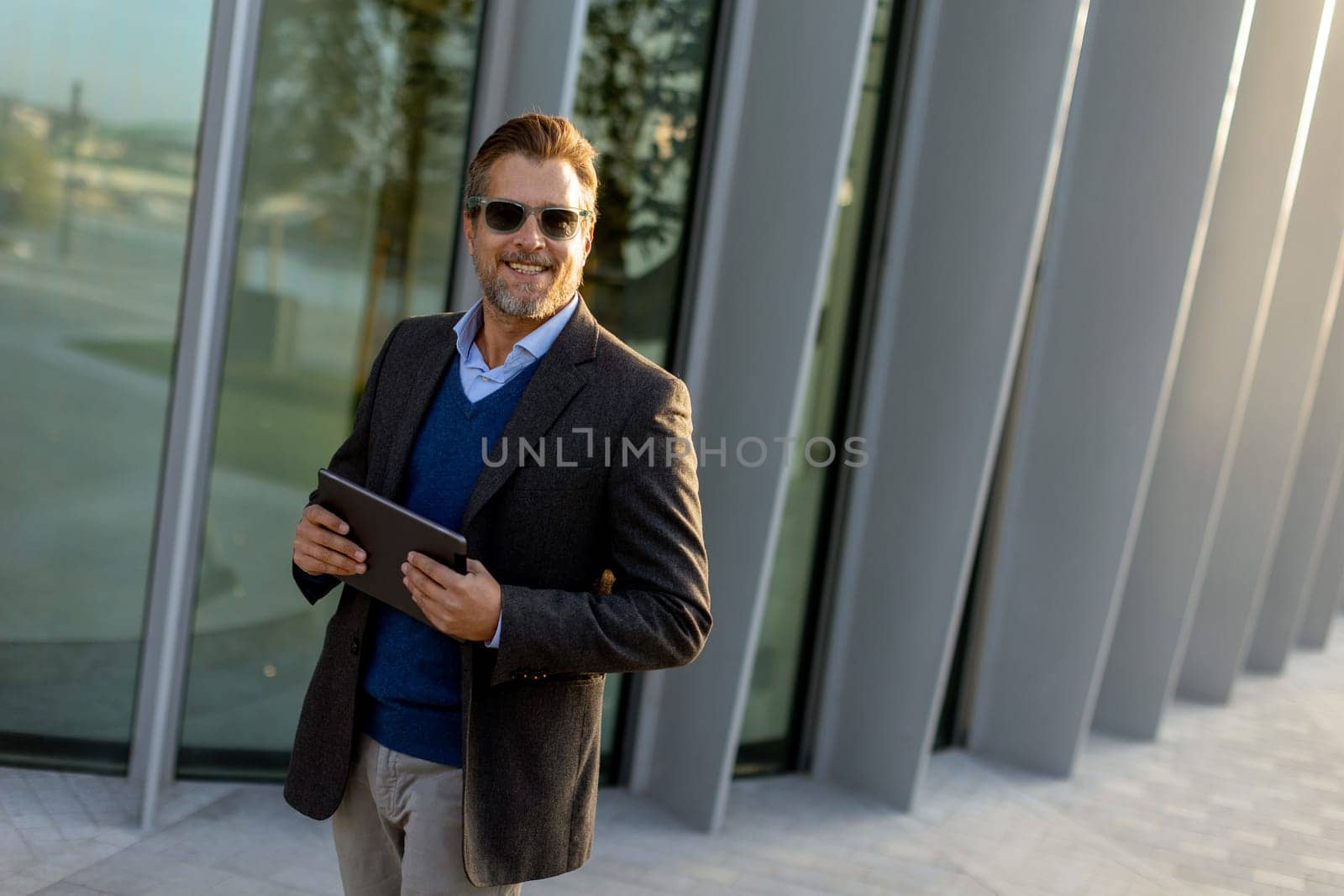A man in business attire stands relaxed against a sleek structure, smiling while holding a tablet, embodying a blend of professionalism and leisure on a sunny day.