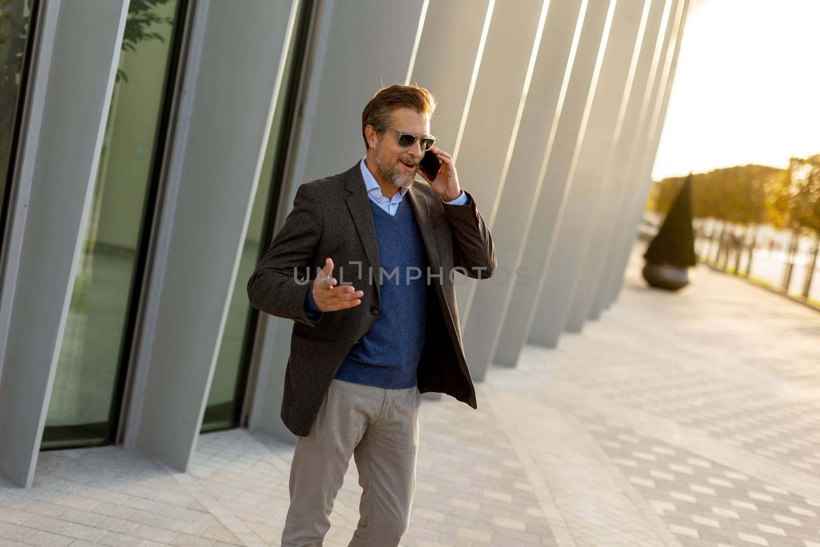 A well-dressed man enjoys a casual conversation on the phone while walking along a sunlit pathway next to contemporary buildings, creating an ambiance of sophistication.