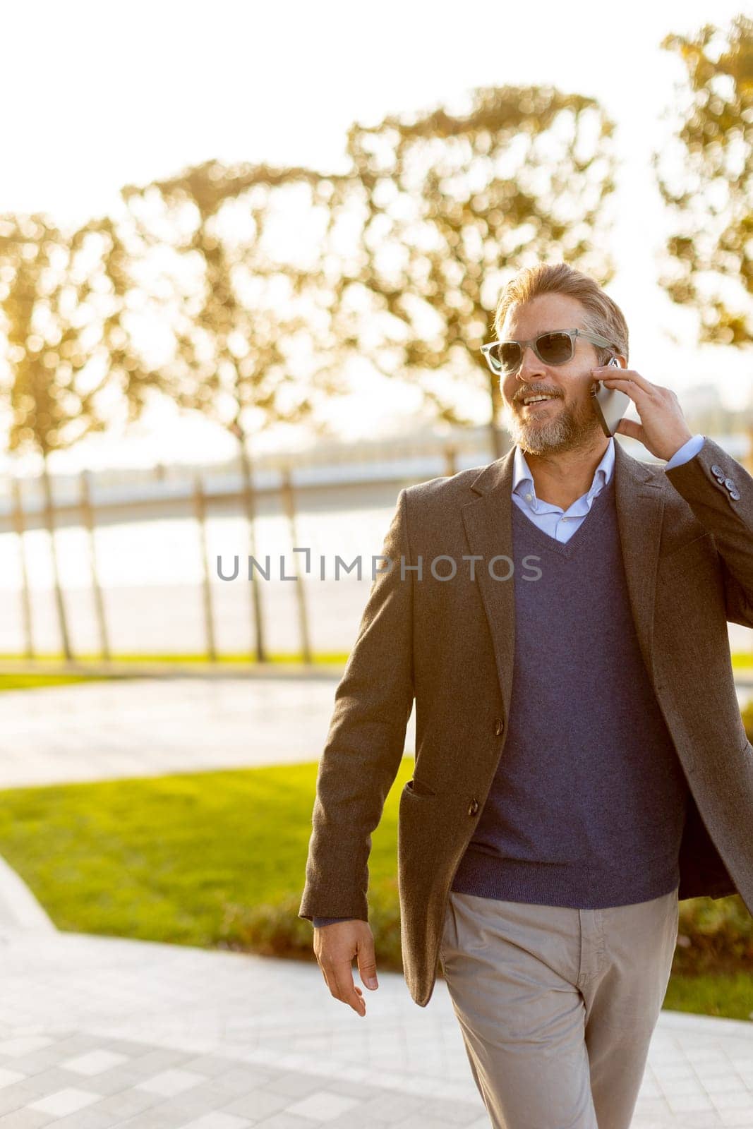 A professional man dressed in smart casual attire is engaged in a phone conversation, smiling as he strolls through a vibrant park. The sun sets, casting a warm glow.