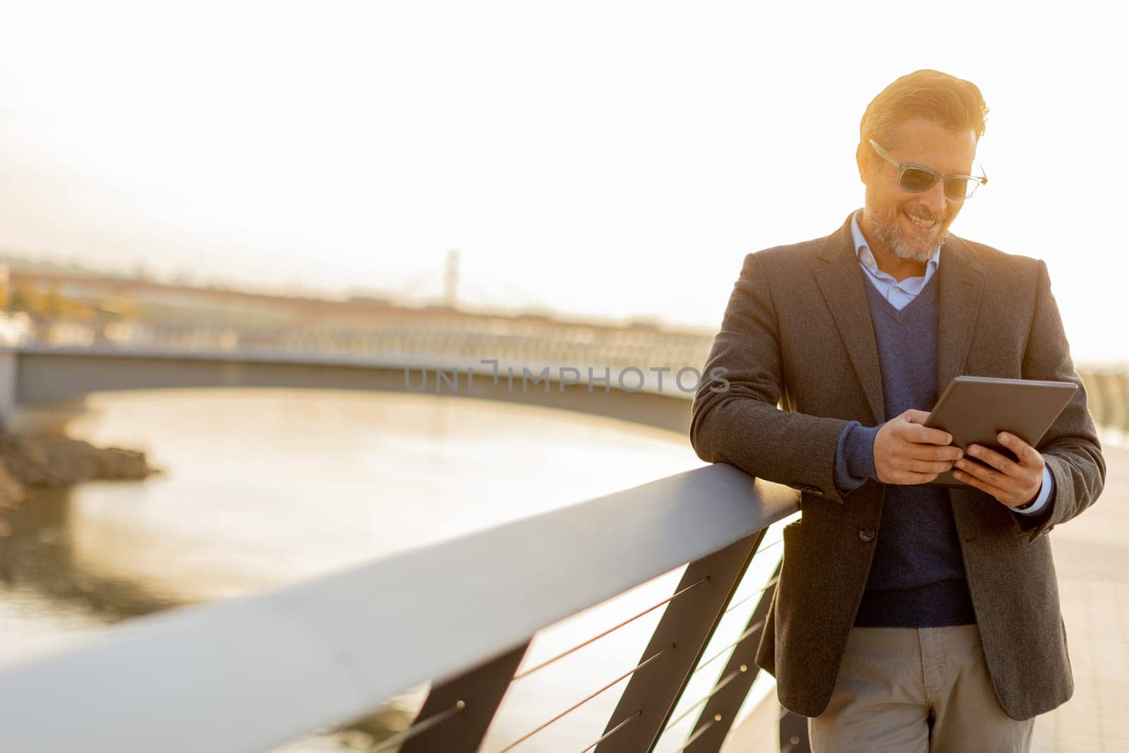 Wearing a stylish outfit, a man with a warm smile leans against a railing by the river at sunset, engaging with his tablet in a peaceful and picturesque environment.