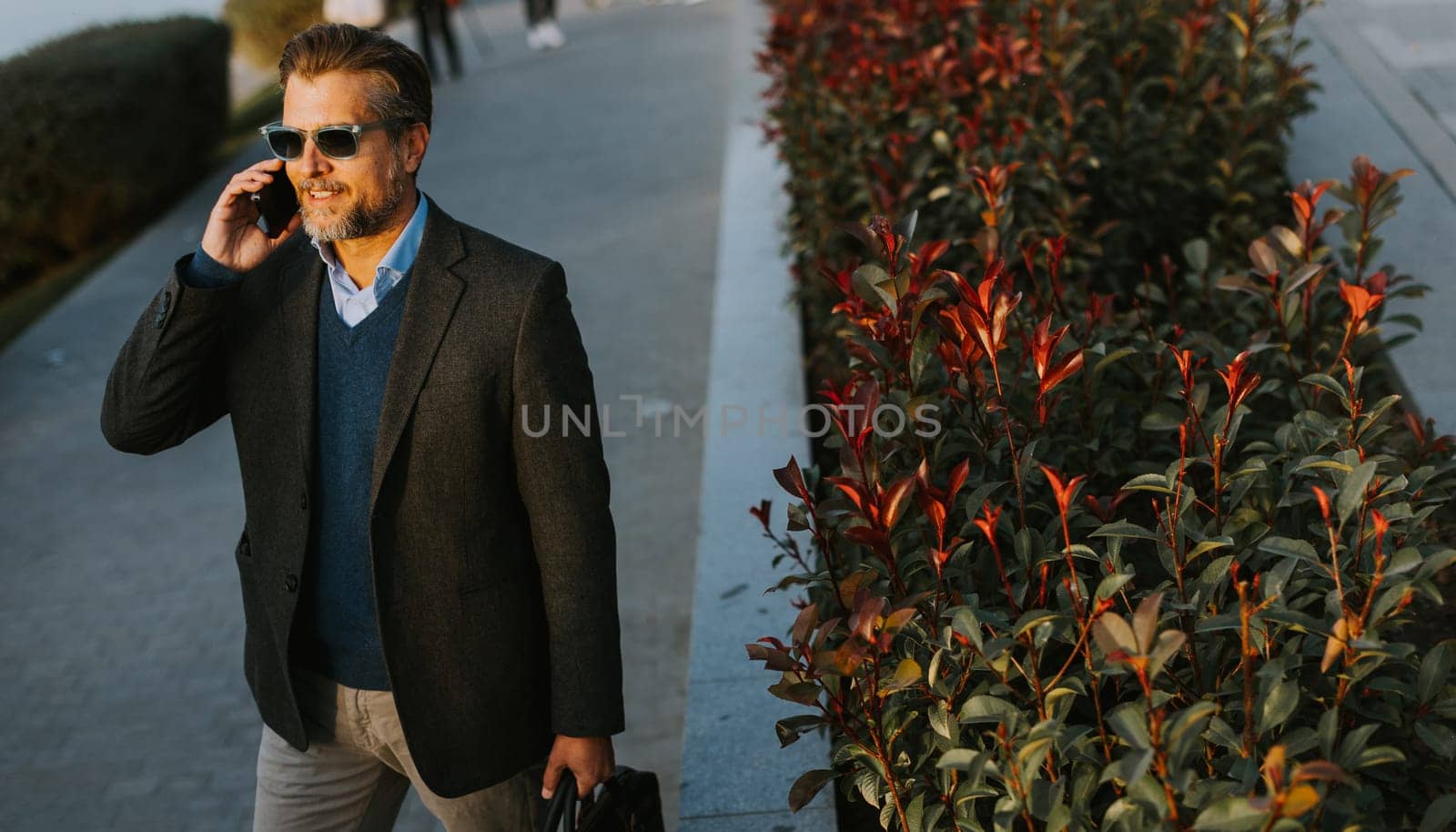 A well-dressed man strolls along a riverside walkway while talking on his phone. The vibrant plants and calming water create a serene atmosphere perfect for business discussions.