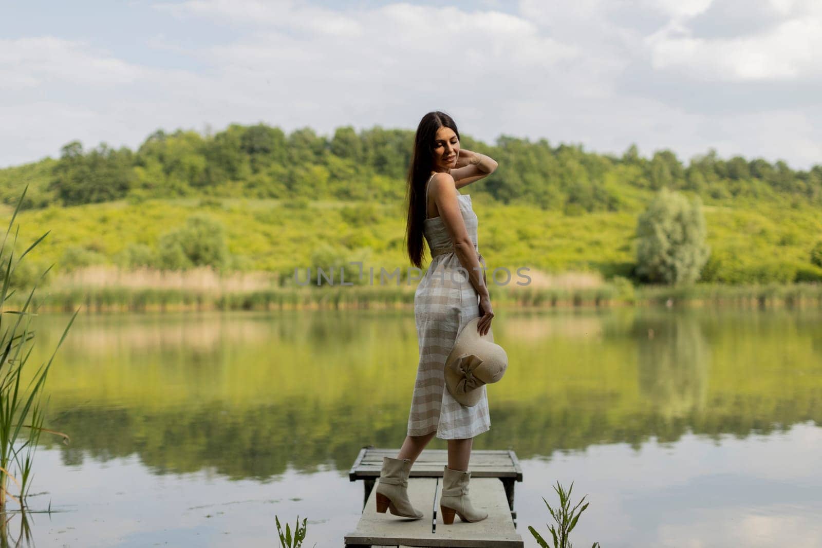 Young woman standing on the wooden pier at the calm lake on a hot summer day