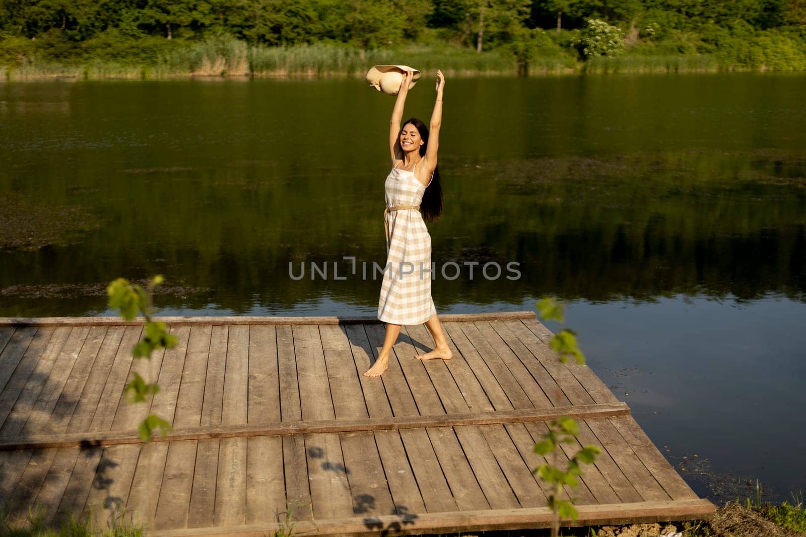 Happy young woman standing on the wooden pier at the calm lake on a hot summer day