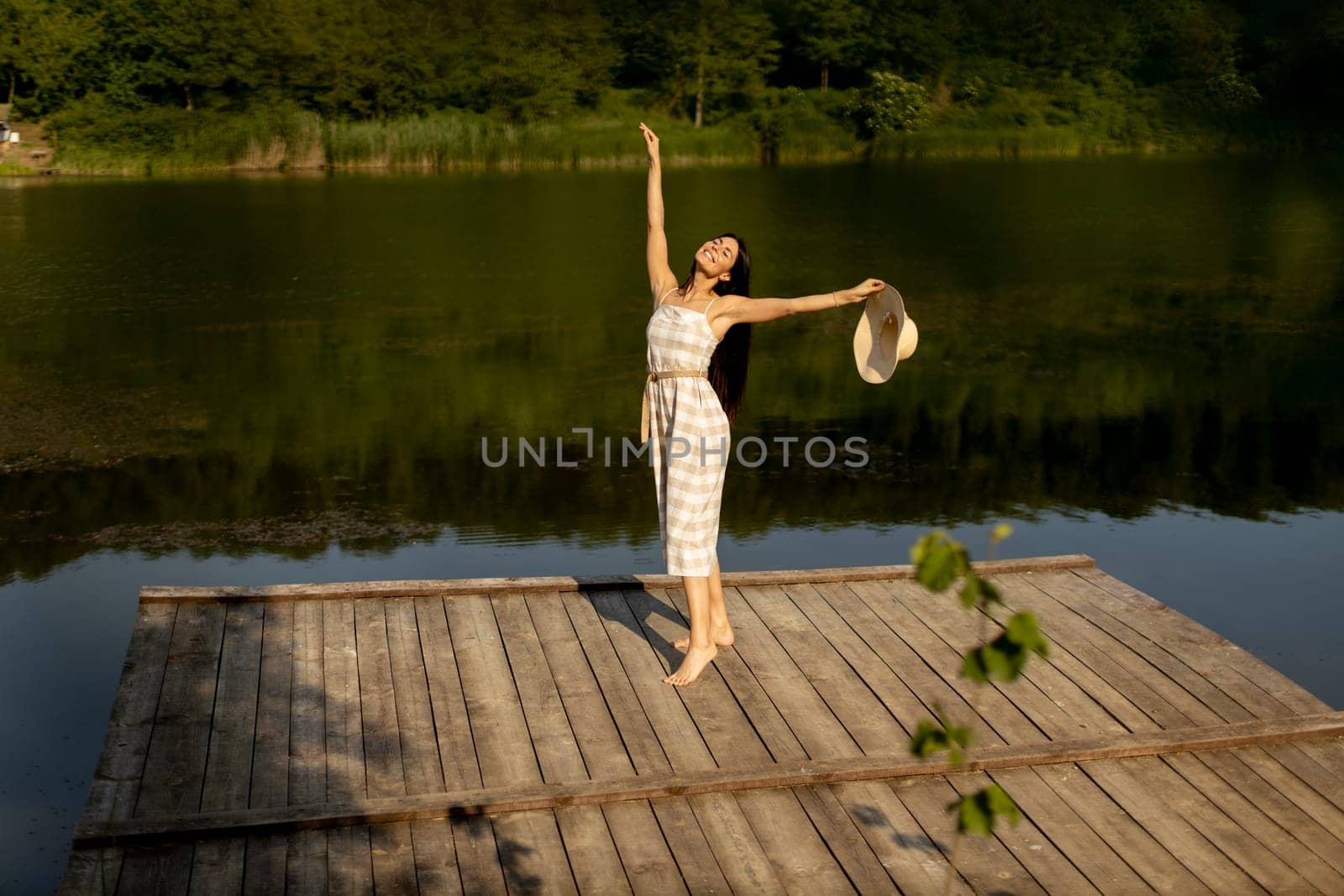 Happy young woman standing on the wooden pier at the calm lake on a hot summer day