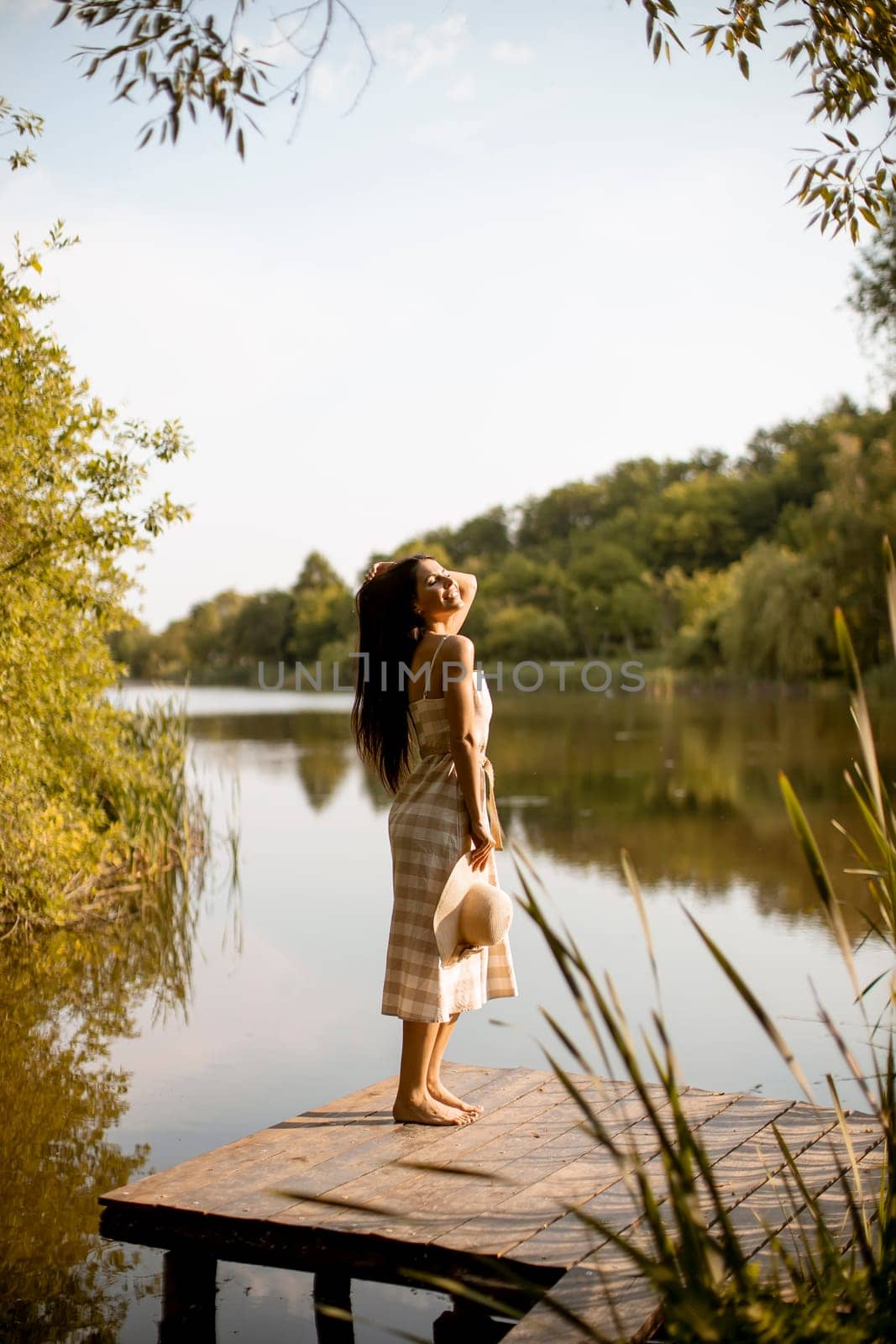 Young woman standing on the wooden pier at the calm lake on a hot summer day