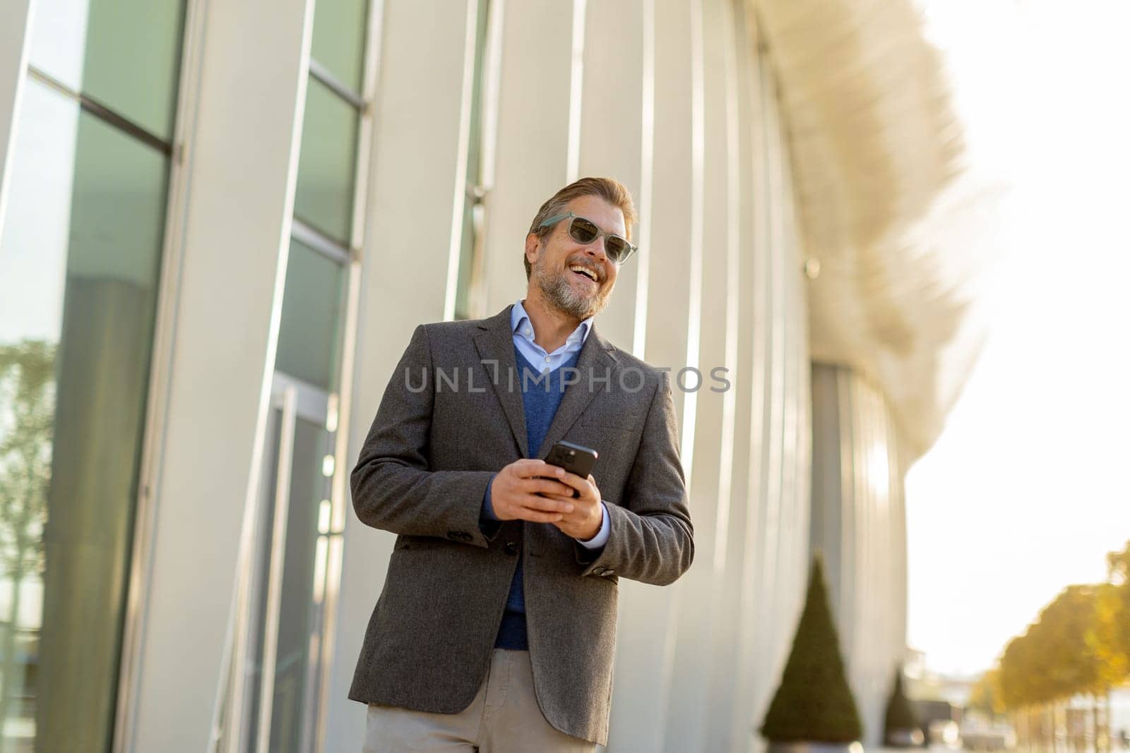 A professional man in stylish attire leisurely walks outside a sleek building, smiling as he interacts with his phone under the warm afternoon sun, exuding confidence.